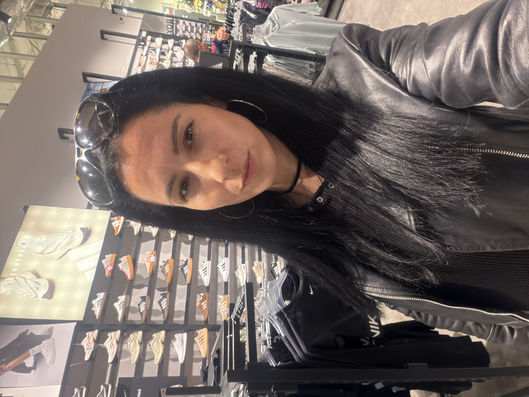 A woman with dark hair, wearing a black leather jacket and sunglasses on her head, is taking a selfie inside a store. Behind her, displays of shoes, including Adidas and Nike brands, are visible. Clothing racks are also present. The specific location and city of Pleven, Bulgaria, cannot be determined from this image.