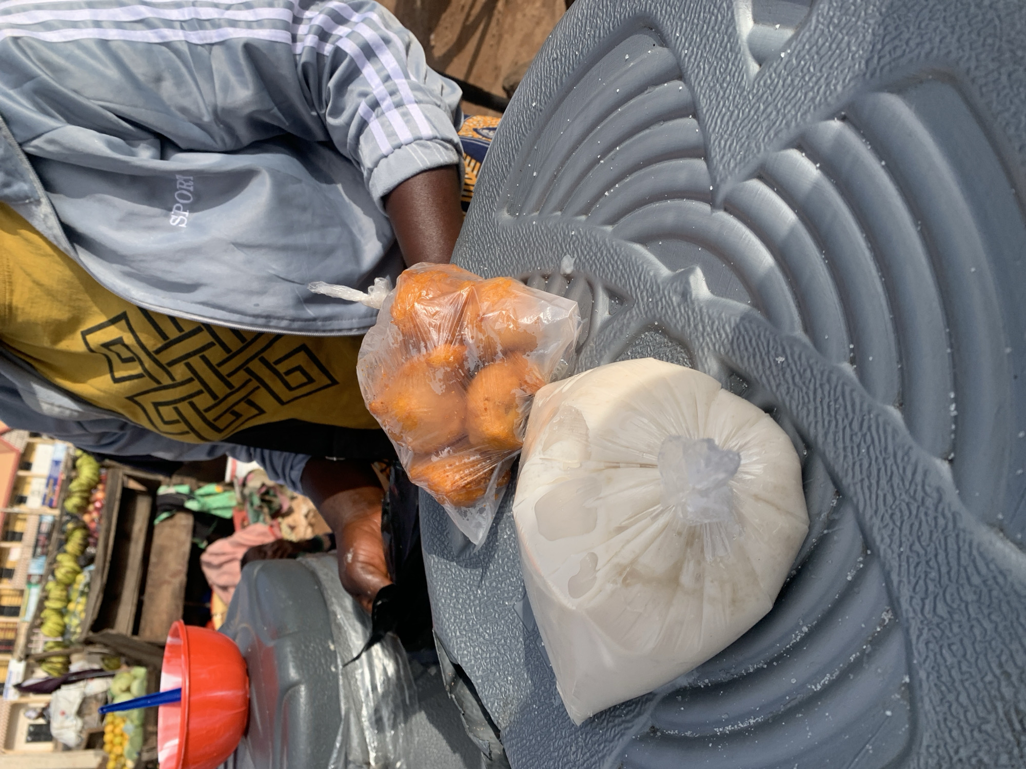 The image depicts a street vendor in Gura Top, Nigeria, attending to food items. In the foreground, two plastic bags are placed on a textured gray surface. One bag contains a creamy white substance, possibly a drink or dairy product, while the other holds several round, orange-colored fritters. Behind these, a person wearing a yellow shirt with a black geometric pattern and a gray jacket with white stripes on the sleeves is visible. The person's hands are shown handling some of the items. To the left, a red bowl with a blue utensil is positioned on a gray container. The background suggests an outdoor market setting, with stalls displaying fruits such as bananas and other produce.