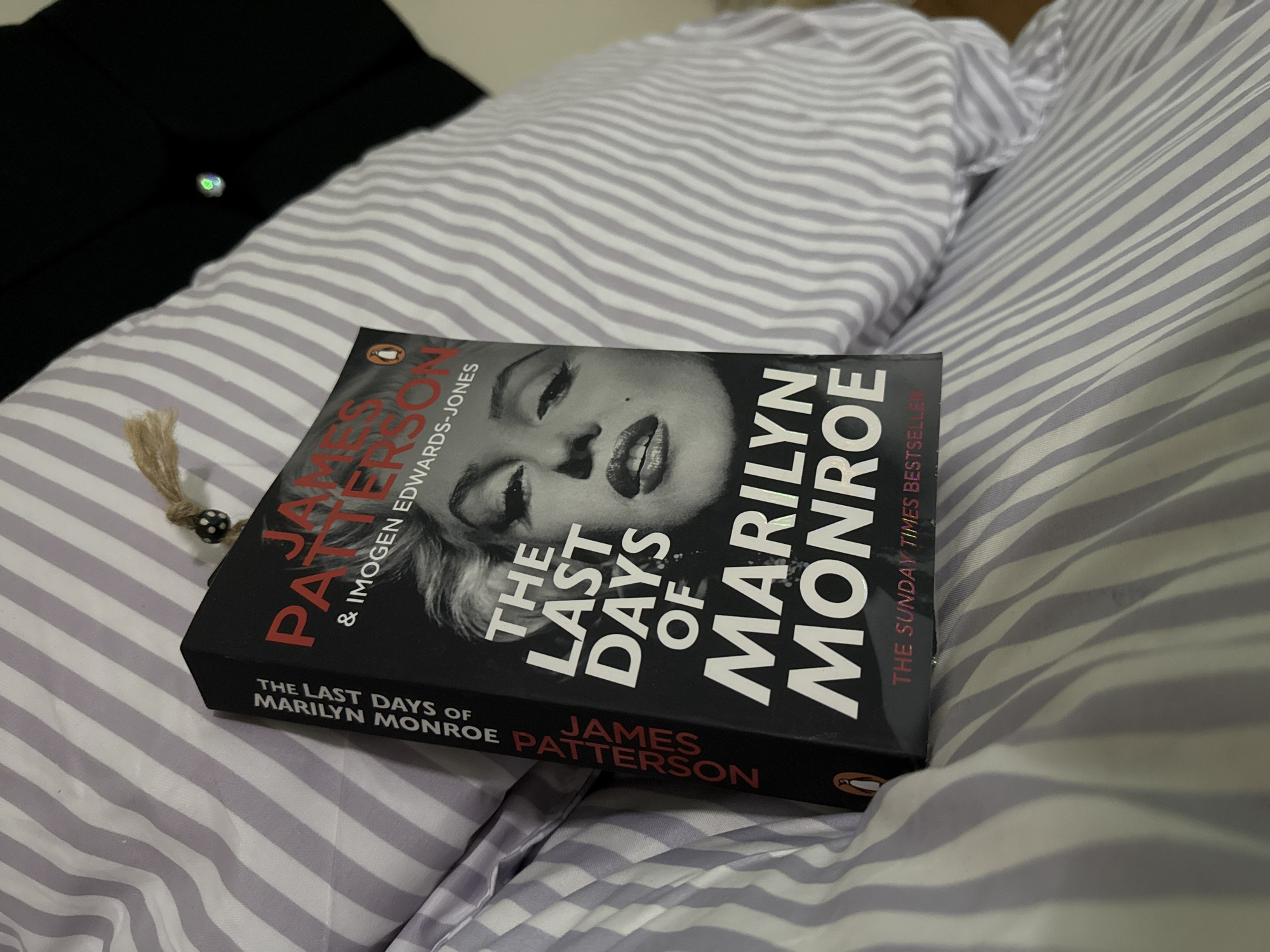 A close-up, eye-level shot captures a paperback book titled "The Last Days of Marilyn Monroe" by James Patterson and Imogen Edwards-Jones, resting on a bed. The book cover is black, featuring a prominent black and white close-up image of Marilyn Monroe's face, with her signature beauty mark visible. The title is in large white sans-serif font, and the authors' names are in red and white. The spine also displays the title and James Patterson's name. A small Penguin Books logo is visible on the bottom right of the cover.
The book is positioned diagonally across a white and light grey (or lavender) striped duvet or bedding. To the left of the book, a small, dark-colored bead with white polka dots is attached to a textured, light-brown string, possibly part of a bookmark, dangling near the book's spine. In the blurred background, a dark, solid-colored item, possibly a garment or another piece of bedding, is visible. The lighting is artificial and dim, suggesting an indoor setting like a bedroom, possibly in the evening. The scene conveys a relaxed atmosphere, perhaps before or after reading. No specific location cues are visible to confirm Great Yarmouth, United Kingdom.