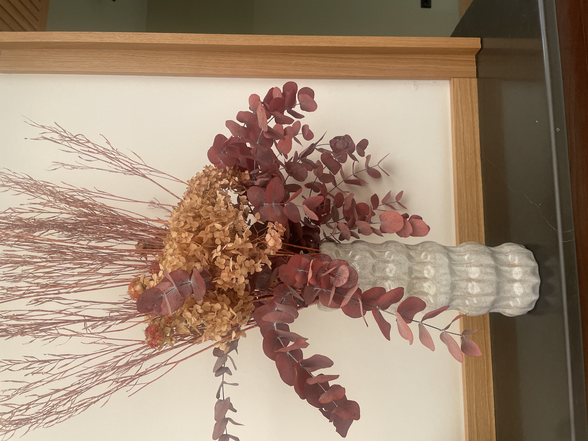 A close-up, eye-level shot captures a decorative vase filled with dried flowers and branches. The vase, made of a textured, light-colored material, has a distinctive ribbed pattern. The floral arrangement includes delicate, reddish-brown branches extending upwards, clusters of small, dried, light brown flowers, and branches with oval, reddish-purple leaves that cascade downwards. The vase rests on a dark, reflective surface, possibly marble or granite, which is set against a light-colored wall. To the right of the arrangement, a vertical wooden frame with a natural finish is visible, suggesting the composition is part of an interior design element. The overall lighting is soft, highlighting the textures and colors of the dried botanicals. The scene is static and does not contain any people or animals. This image does not provide any specific details or context that would allow for its location to be identified as Sevilla, Spain.