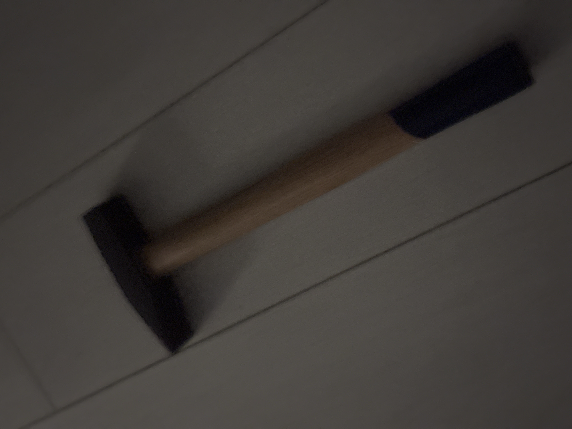 A single, blunt-headed hammer with a wooden handle and a dark blue grip lies diagonally across a light-colored, linoleum floor. The hammer is positioned from the upper left to the lower right of the frame. The floor appears to be made of planks, with visible lines suggesting seams between them. The overall lighting is dim, casting long shadows and giving the scene a muted, somber tone. There are no people or any other objects visible in the image. The location is not identifiable as Sofia, Bulgaria.