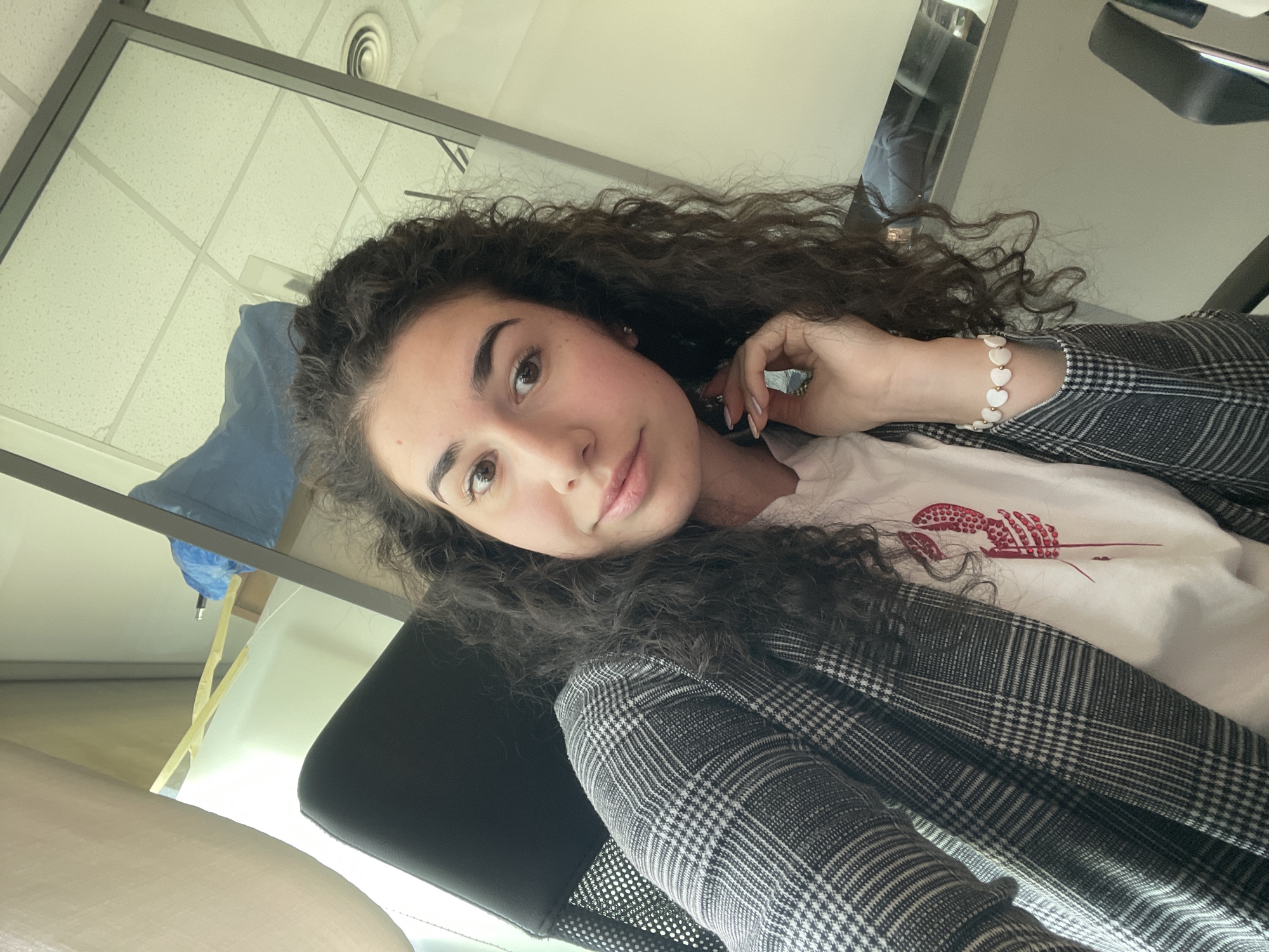 A young woman with curly, dark hair poses for a selfie indoors. She is wearing a gray and white checkered blazer over a white t-shirt with a red design. Her left hand is raised near her face and adorned with a beaded bracelet. The background features an office setting with a drop ceiling, windows, and office furniture. The location cannot be determined to be in the city of Sofia, Bulgaria from the image.
