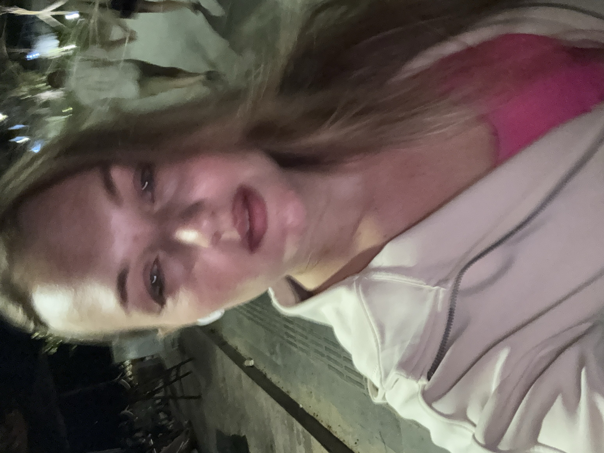 A woman with long blonde hair, wearing a pink top and a light-colored zippered jacket, takes a selfie at night. She has on white wireless earbuds. In the background, the street is illuminated by lights, and other people can be seen walking. The scene appears to be outdoors in a public area, possibly a street or a walkway. The location is a street in Denpasar, Indonesia.