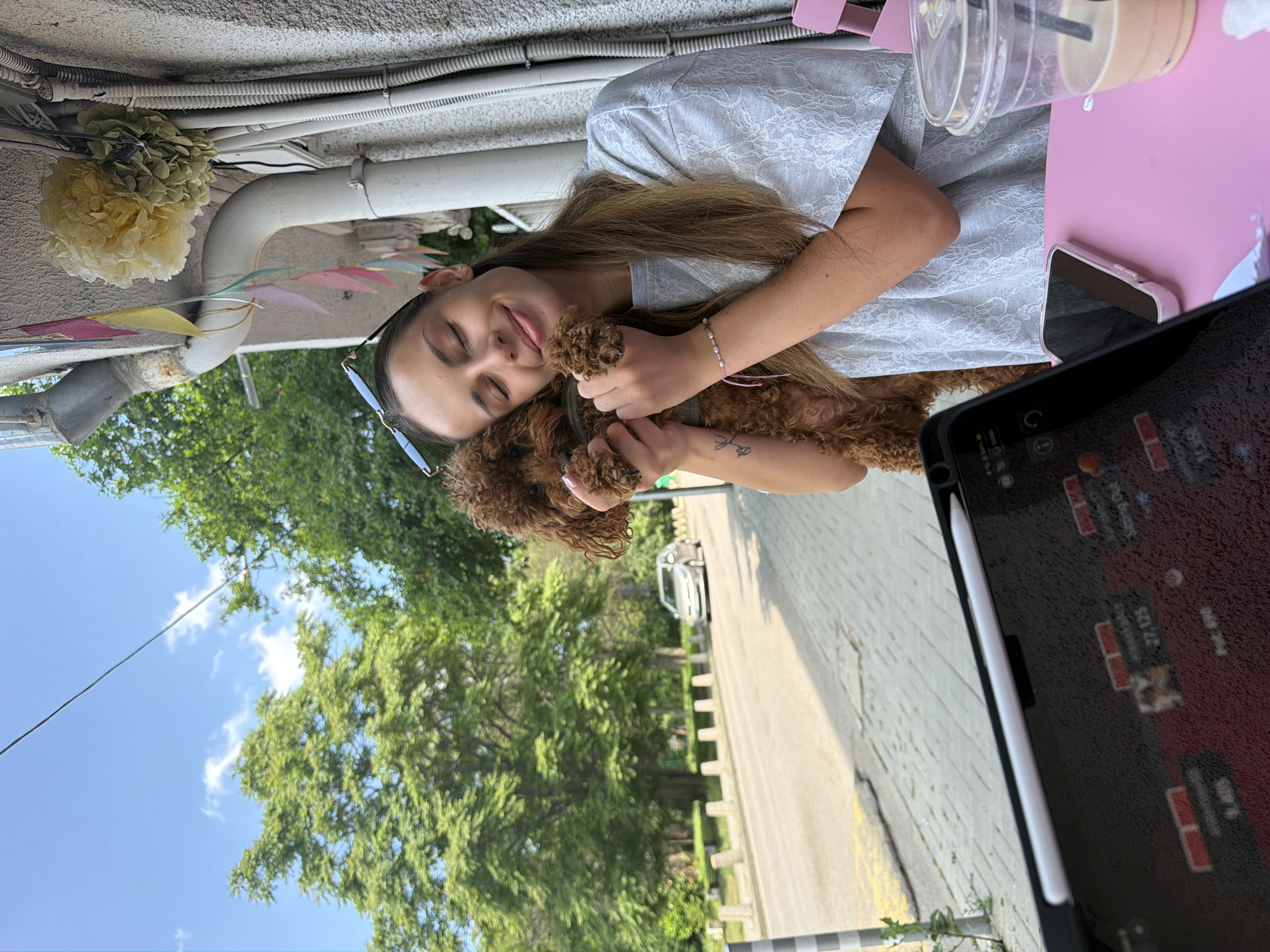 The scene unfolds outdoors in the city of Sofia, Bulgaria, depicting a young woman affectionately holding a small, curly-haired brown dog against her chest. The woman, with long brown hair and sunglasses resting on her head, wears a light grey patterned short-sleeved top and has a pink beaded bracelet on her right wrist, with a small tattoo on her left forearm. Her eyes are closed, and she smiles gently, indicating contentment.
She is seated at a pink outdoor table, where a dark-cased tablet with a white stylus is positioned vertically, displaying what appears to be game or poker statistics with numbers such as "27,125". A pink-cased smartphone lies face down next to it. To the right, a plastic cup with a straw contains a light brown beverage.
The background features a light-colored, textured building wall adorned with colorful paper bunting flags and two large decorative pom-poms (one yellow, one pale green). A weathered metal downspout and various white conduits are visible on the wall. To the left, a paved street is visible, with a silver car driving in the distance. The street edge is marked by a black and white striped pole and several low, light-colored bollards. Dense green trees line the street, suggesting a park or tree-lined boulevard, under a clear blue sky with scattered white clouds.