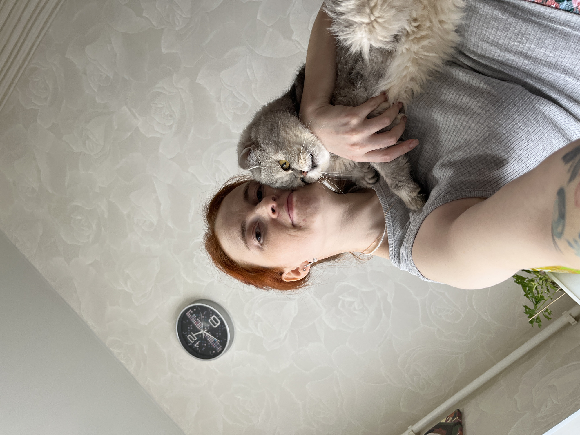 A woman with red hair is holding a gray cat. The woman is wearing a gray tank top and a silver necklace. She is facing the camera. The cat is resting on her shoulder, being held by the woman's hand, with its head next to the woman's face. Behind the woman is a wall with a floral pattern. To the left of the woman's head is a round wall clock. The woman's arm is visible on the right of the image. There is a plant and part of a shoe also visible near the woman's arm. The photograph was likely taken indoors in Chaikovsky, Russia.