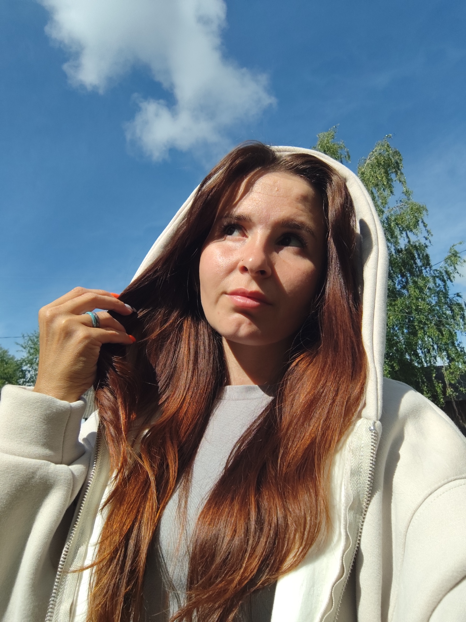 A medium close-up outdoor portrait shows a woman with long, wavy reddish-brown hair, looking upwards and slightly to her right. She wears a light-colored, possibly off-white, hooded jacket with the hood pulled up, over a lighter grey t-shirt. Her left hand is raised, gently touching strands of her hair near her ear. She has a light blue ring on her left ring finger and her fingernails are painted orange. A small nose piercing is visible on her left nostril.
The scene is brightly lit by direct sunlight, casting strong highlights on her forehead, nose, and chin, and creating shadows that accentuate her features. The background is a vivid blue sky with scattered white, fluffy clouds. On the right side of the frame, the green leaves of a tree are visible against the sky. The image suggests a sunny daytime setting, likely outdoors in a natural or semi-natural environment, potentially in Фоки, Russia, based on the provided context. The woman's expression appears contemplative or observant as she gazes skyward. No text is visible in the image.