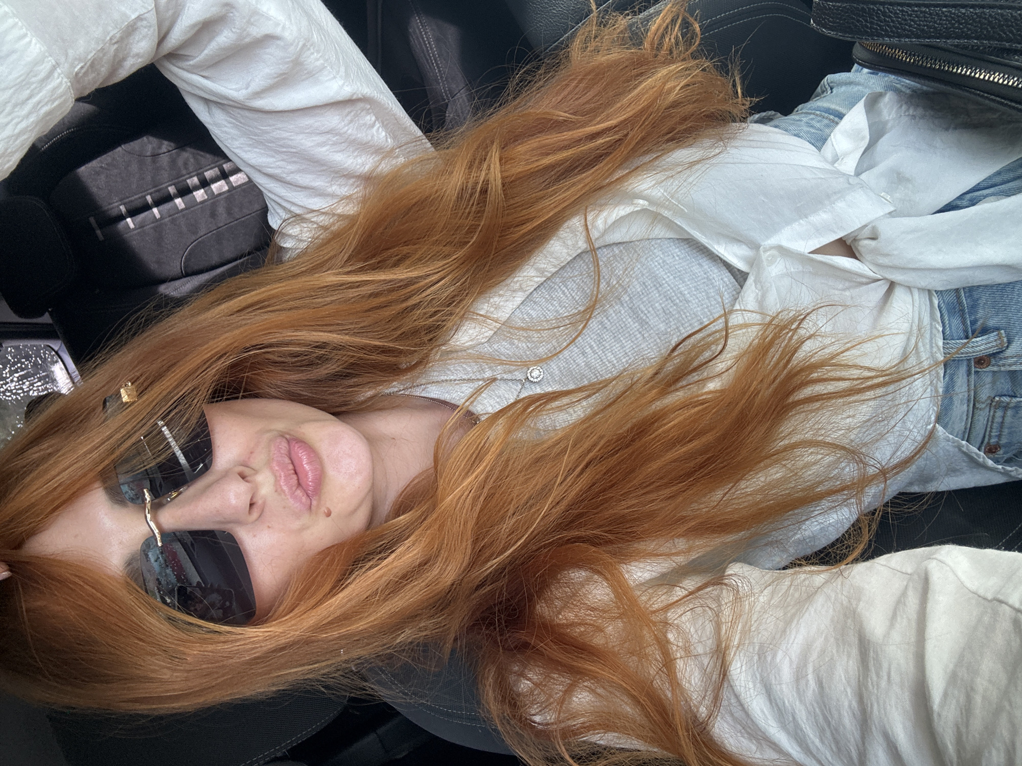 A person with long, flowing, bright reddish-brown hair is captured in a high-angle, medium close-up shot, lying down and looking towards the camera. The individual wears large, dark, rectangular sunglasses with gold-toned accents on the frames, obscuring their eyes. Their lips are slightly parted in a relaxed manner, and a small mole is visible above their upper lip on the right side.
They are dressed in a loose white button-up shirt worn open over a light grey top, with blue denim jeans visible below. A thin silver necklace featuring a small, circular, possibly jeweled pendant rests on their chest.
The setting appears to be the interior of a vehicle, likely a car, indicated by the black upholstered seats surrounding the person. On the left, a black surface with a subtle checkered or striped pattern, consistent with car seat upholstery, is visible. A black bag with a silver zipper can be seen in the upper right corner. The scene is brightly lit, suggesting daytime conditions. The overall impression is one of relaxation. The given location context for this image is Budva, Montenegro. There is no visible text in the image.