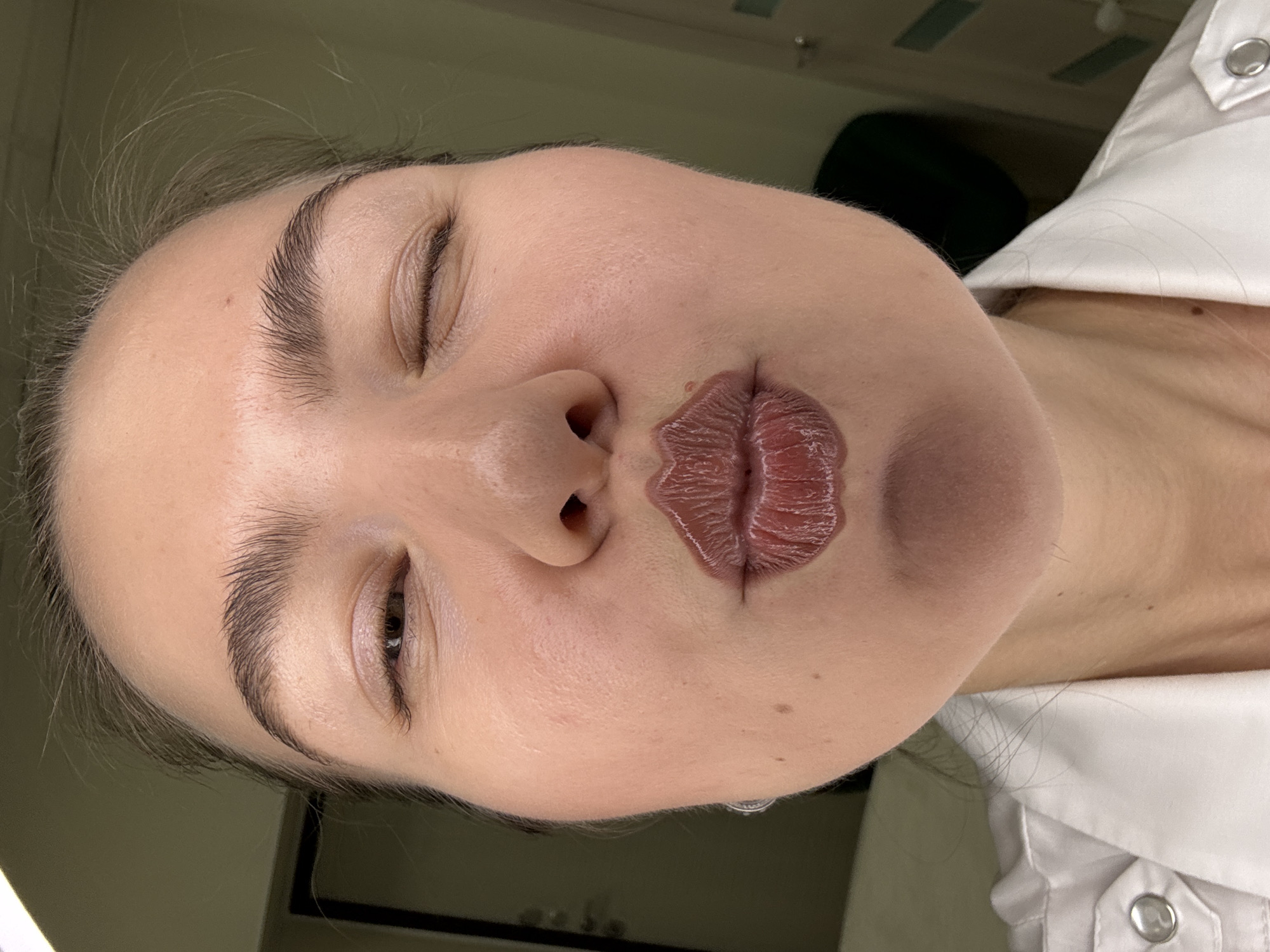 A close-up, front-facing shot shows a woman puckering her lips in a kiss. One eye is open, and the other is closed in a wink. She is wearing a white collared shirt with silver buttons. Her lips are covered in a glossy, dark red lipstick. Her eyebrows are thick and well-defined. The background is blurry, with a hint of a doorway and a green wall. The lighting is soft and even. The image is taken indoors in an unknown location.