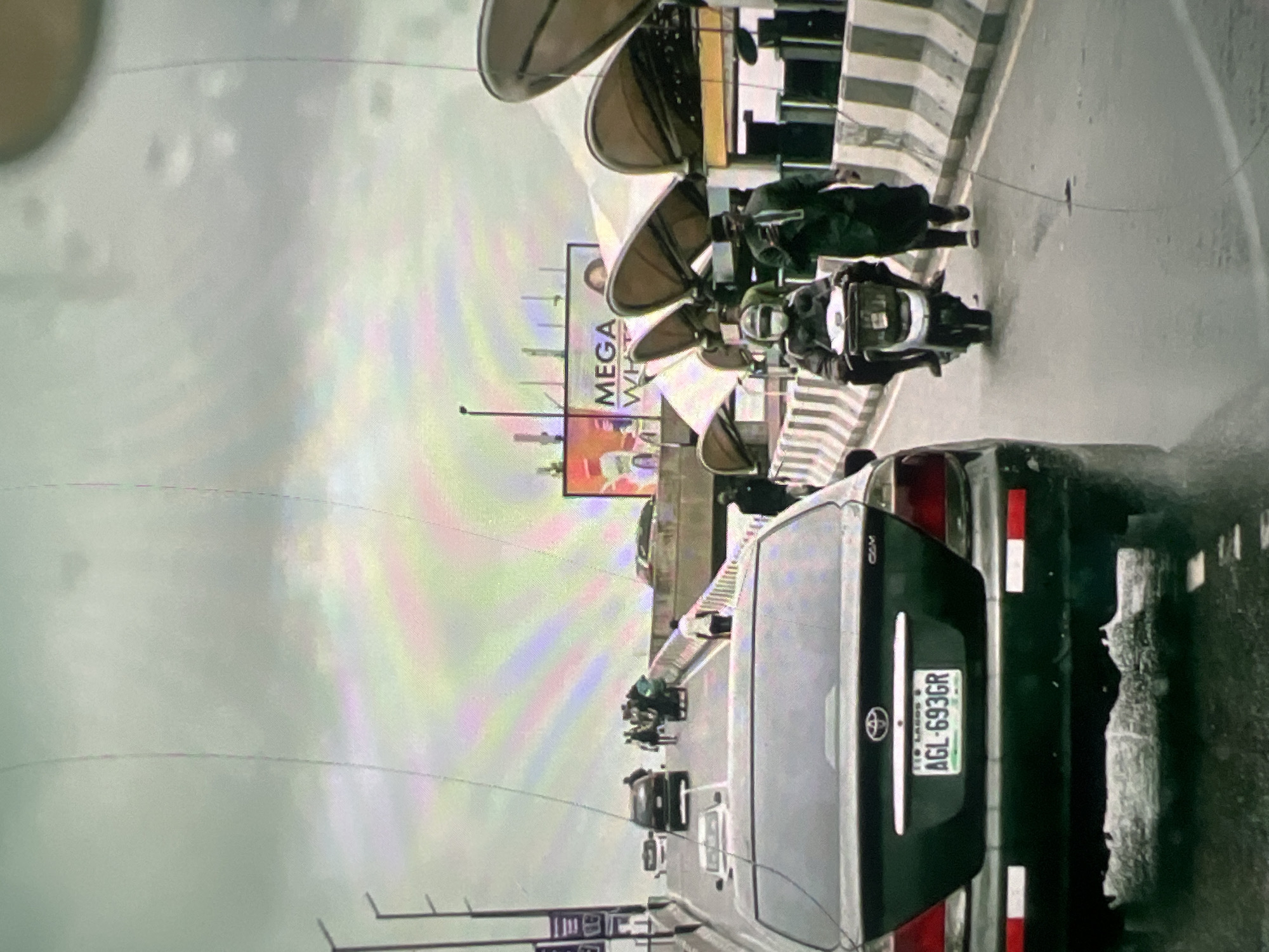 The image depicts a street scene in Lagos, Nigeria, likely on an overpass or elevated road given the presence of multiple lanes and concrete barriers. The sky is overcast with a visible rainbow-like atmospheric effect.
In the foreground, the rear of a dark-colored Toyota sedan is prominently featured, displaying the license plate "AGL-693GR" from Lagos. To the right of this vehicle, a person on a scooter is seen wearing a mask and helmet. Further ahead, another individual in a green traditional outfit and cap is walking and looking at a smartphone.
In the background, traffic is visible, including several cars and a group of pedestrians crossing the road. A large billboard advertising "MEGA WHITE" is also present. The road surface appears wet, suggesting recent rain. The overall impression is a busy urban environment under a dramatic sky.