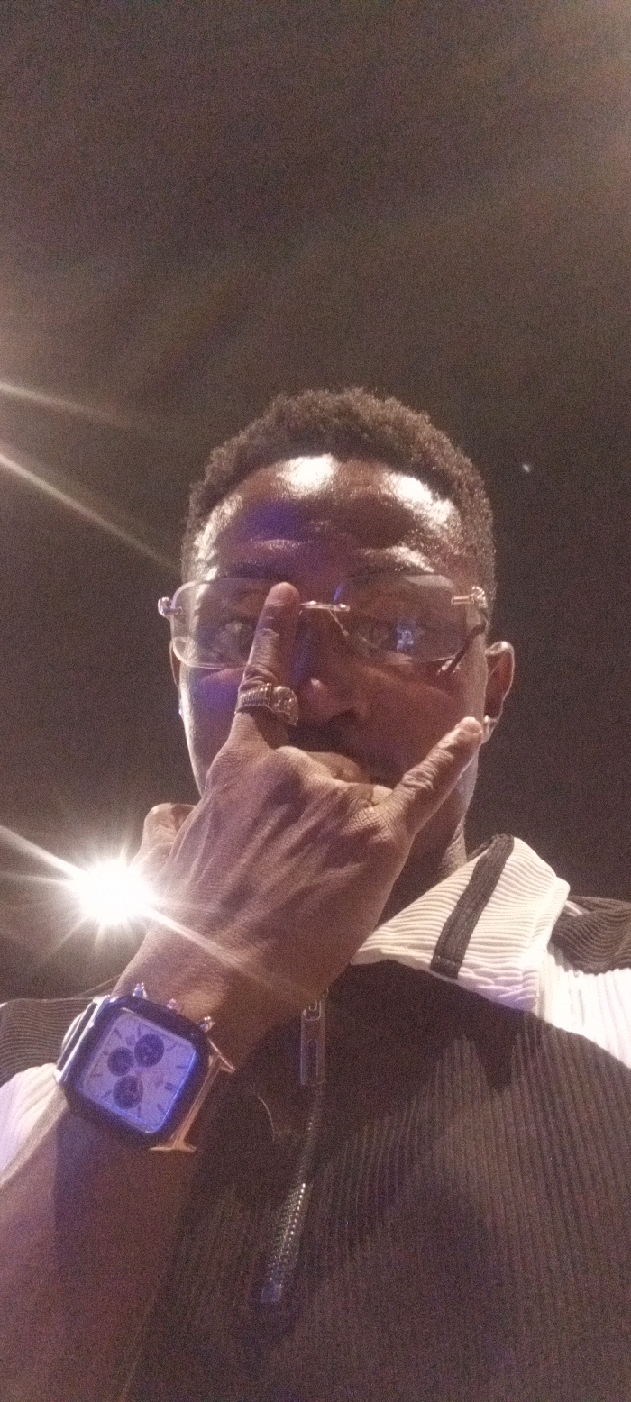 The image presents a man, likely in Lokoja, Nigeria, captured in a dimly lit environment, suggesting either an indoor setting or nighttime. He is positioned from the chest up, looking towards the camera with a confident pose.
The man has short, dark, curly hair and is wearing clear-rimmed or thinly-framed glasses that reflect ambient light. A large, ornate gold-toned ring adorns the index finger of his right hand. On his left wrist, he wears a prominent square-faced watch with a rose gold or copper-colored casing and a dark strap. The watch face displays multiple dials or chronographs, illuminated with a bluish-purple light.
He is dressed in a black top featuring vertical pleats or ribs, with a visible zipper down the front and a white collar accented with a black stripe. His right hand is raised, forming a gesture with the index finger and thumb extended while the other fingers are curled, often recognized as a "rock on" sign.
The background is dark and features several bright light sources on the left side and in the upper left corner, creating noticeable lens flares and reflections. The image quality shows some grain, characteristic of low-light photography. No visible text is discernible on his attire or accessories.