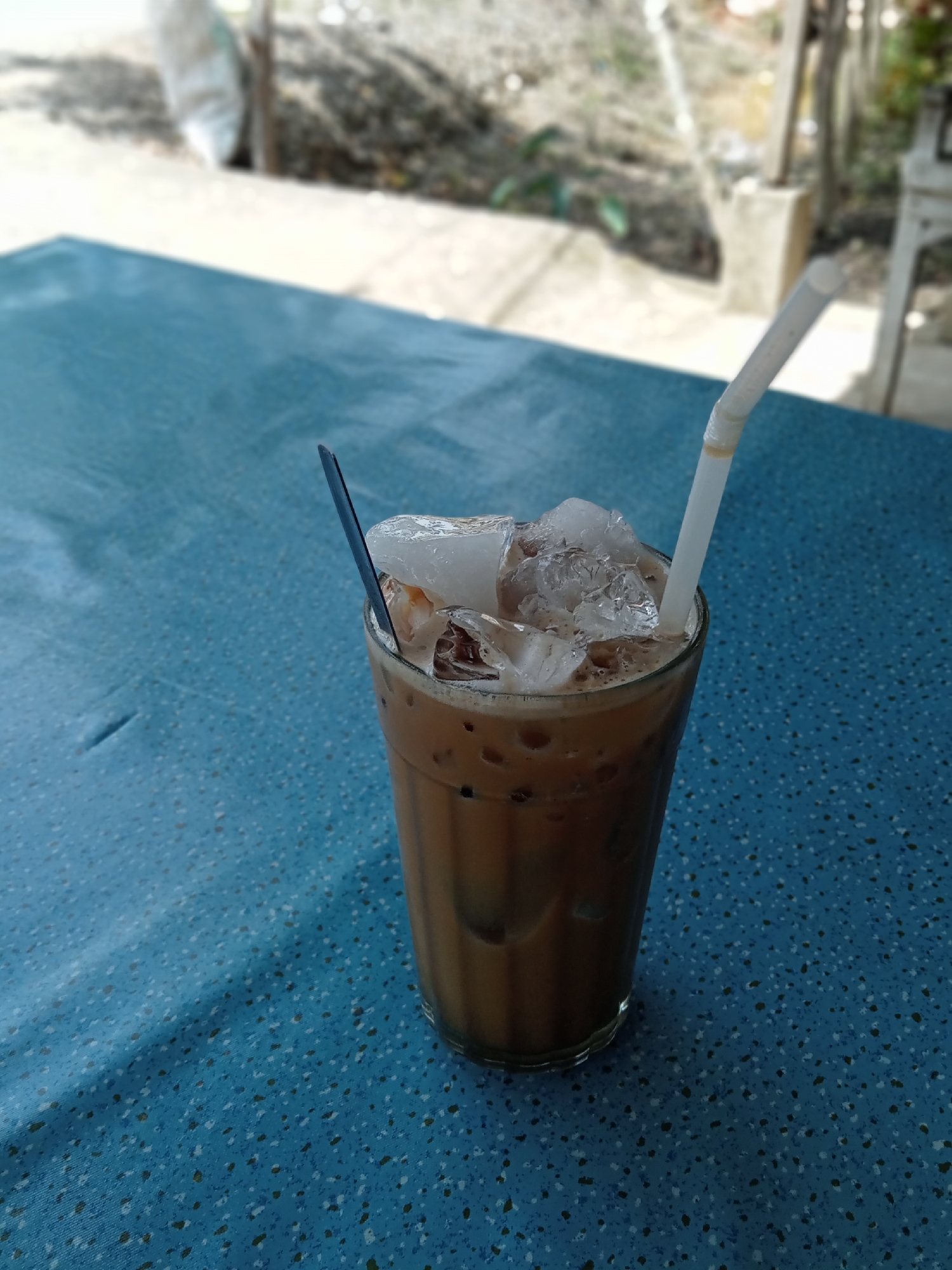 A tall glass of iced coffee, topped with ice cubes and a white straw, sits on a blue, speckled table. A dark spoon is partially submerged in the ice. The background is blurred, showing a hint of outdoor seating and foliage, suggesting a cafe or restaurant setting. Sunlight casts a shadow on the left side of the table. The image appears to be taken from a slightly elevated angle.