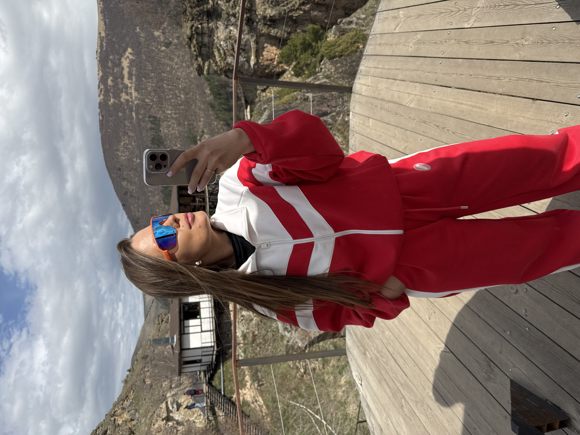 A woman is taking a selfie while standing on a wooden platform in what appears to be a mountainous region. She is wearing a red and white tracksuit and reflective sunglasses. She is holding a smartphone in her right hand. In the background, there is a steep mountain and a building perched on the hillside with a staircase leading to it. The sky is partly cloudy. The location is Unknown, Russia.