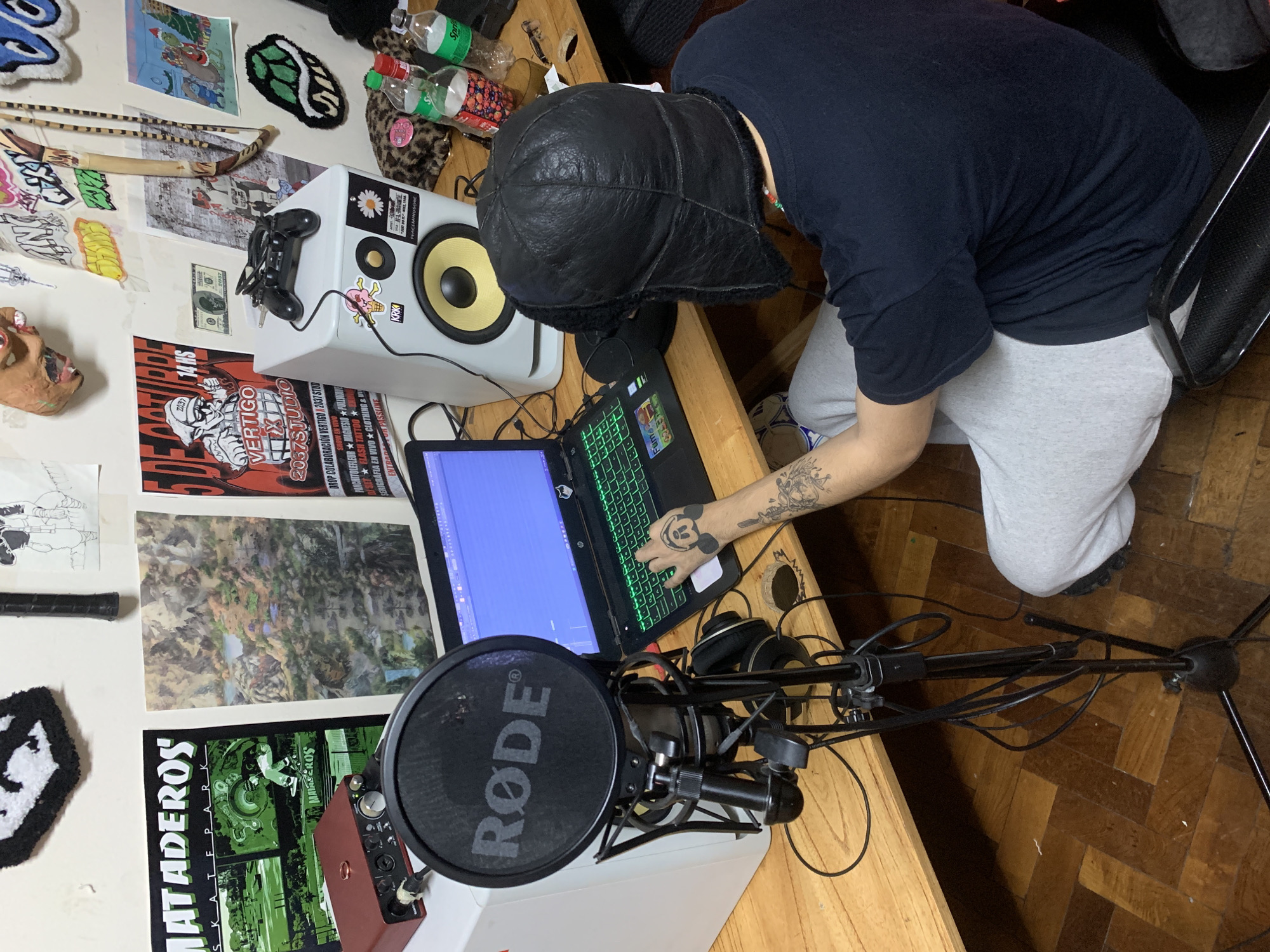 A person wearing a black fur-lined aviator hat, a black shirt, and grey sweatpants sits at a wooden desk. They are operating a laptop with a green keyboard backlight. The person has tattoos on their arm. A Rode microphone with a pop filter is positioned in front of the laptop. A studio monitor speaker is to the left of the laptop, near some small bottles and a game controller. The wall behind the desk is decorated with various posters, stickers, and decorative items including a Mataderos skatepark poster, a Vertigo 2057 Studio poster, a dollar bill, and a small mask. The floor is wooden with a herringbone pattern. The scene is likely a home recording studio. The Mataderos poster suggests a location in the Mataderos neighborhood of Buenos Aires, Argentina.