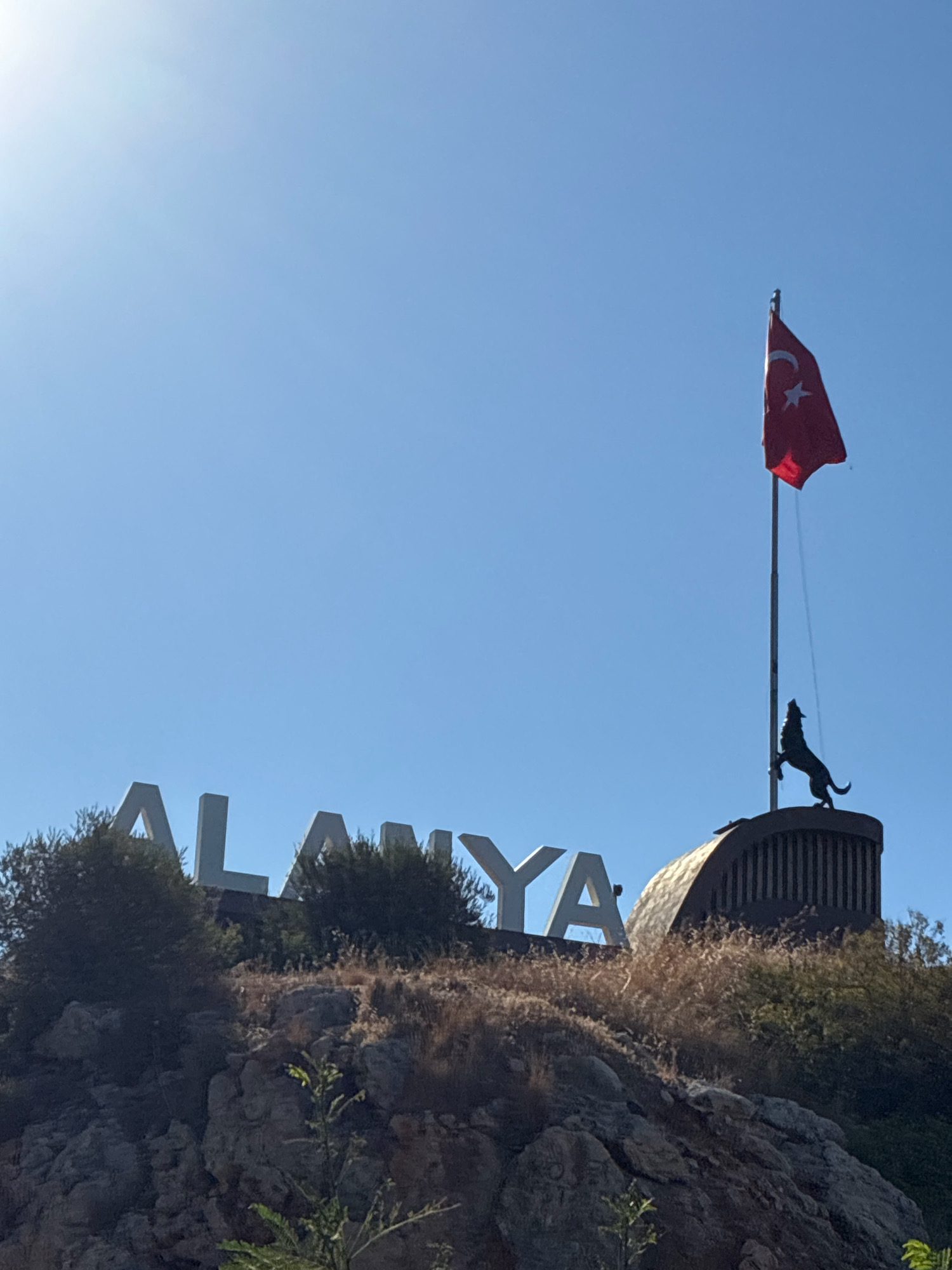 An outdoor, low-angle, full shot captures a bright, sunny day in Alanya, Turkey. The sky is a clear, vibrant blue, with a slight glare in the upper left quadrant indicating the sun's position. In the middle ground, a prominent sign spells out "ALANYA" in large, white, three-dimensional letters, partially obscured by the surrounding foliage and rocky terrain.
To the right of the "ALANYA" sign, on a curved, dark structure, a Turkish flag with a white crescent and star on a red background flies from a flagpole. Below the flag, silhouetted against the sky, is the figure of a wolf in a howling pose, standing on its hind legs. The wolf sculpture is positioned atop a dark, ribbed cylindrical base.
The foreground is dominated by a rugged, rocky hillside covered with dry, golden-brown grasses and scattered green bushes. A few small green plants are visible in the lower part of the frame. The overall impression is of a scenic overlook or monument in Alanya.