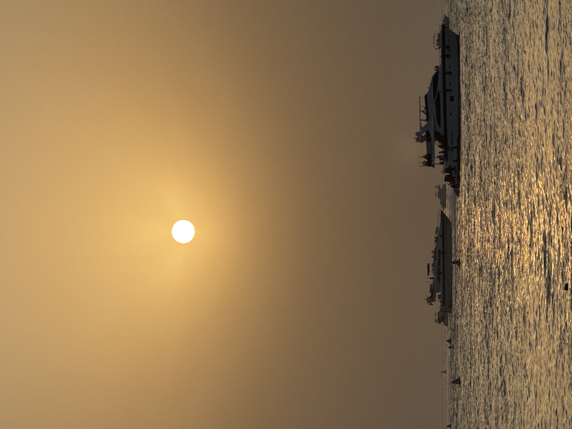 A bright, white sun is centered in the upper half of the frame against a hazy, orange sky. Below, the ocean is depicted with moderate ripples reflecting the golden sunlight. Several white yachts of varying sizes are scattered across the water. One large yacht, centrally located in the lower half of the image, is populated with numerous people. Another, smaller yacht is visible further back and to the left, also with people on board. Several buoys are also present in the water. The scene appears to be a daytime view in Dubai, United Arab Emirates, likely a coastal area.
