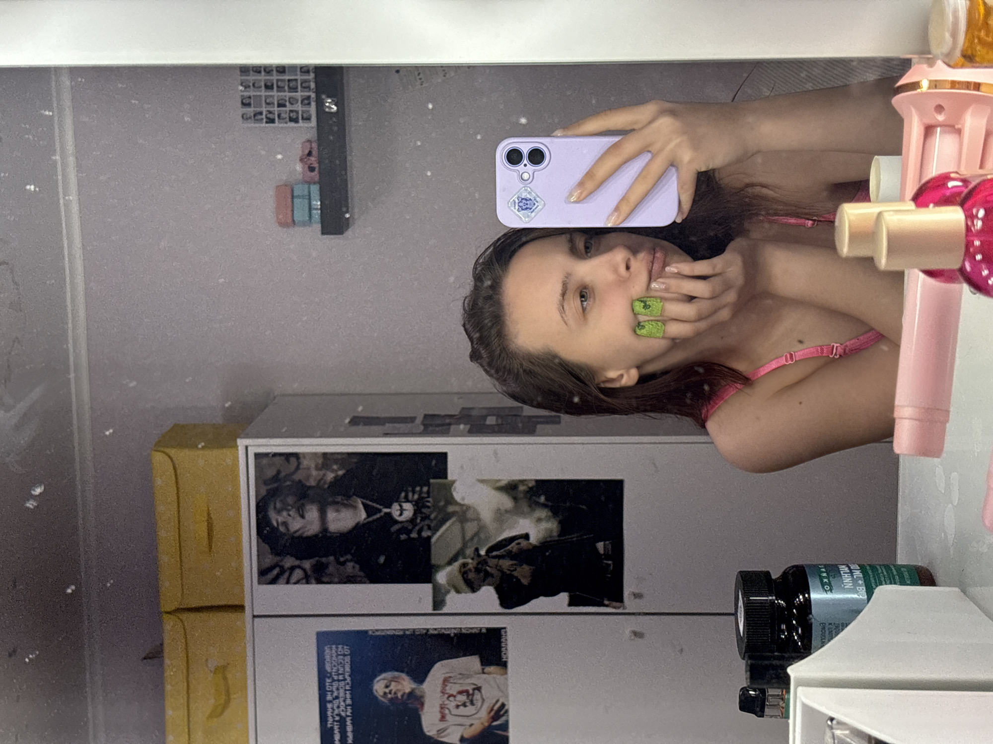 The image is a mirror selfie of a woman with brown hair. She is holding a purple iPhone with a sticker on the back. She is wearing a pink bra and has green nail polish on her fingers. In the background, there is a yellow box, a white cabinet with black and white posters on it, and a black bottle with a green label that says "WL+B", "WYLHNN", "JYNKO", "ENOVOLNA", "10 ПОВЕЪРСИ ИНЕ НУ ЕНРИ", "ОСВОЕР-310 НЕ ЗИМИНЬ". There is also a pink hair accessory on the right side of the image. The location is not discernible.