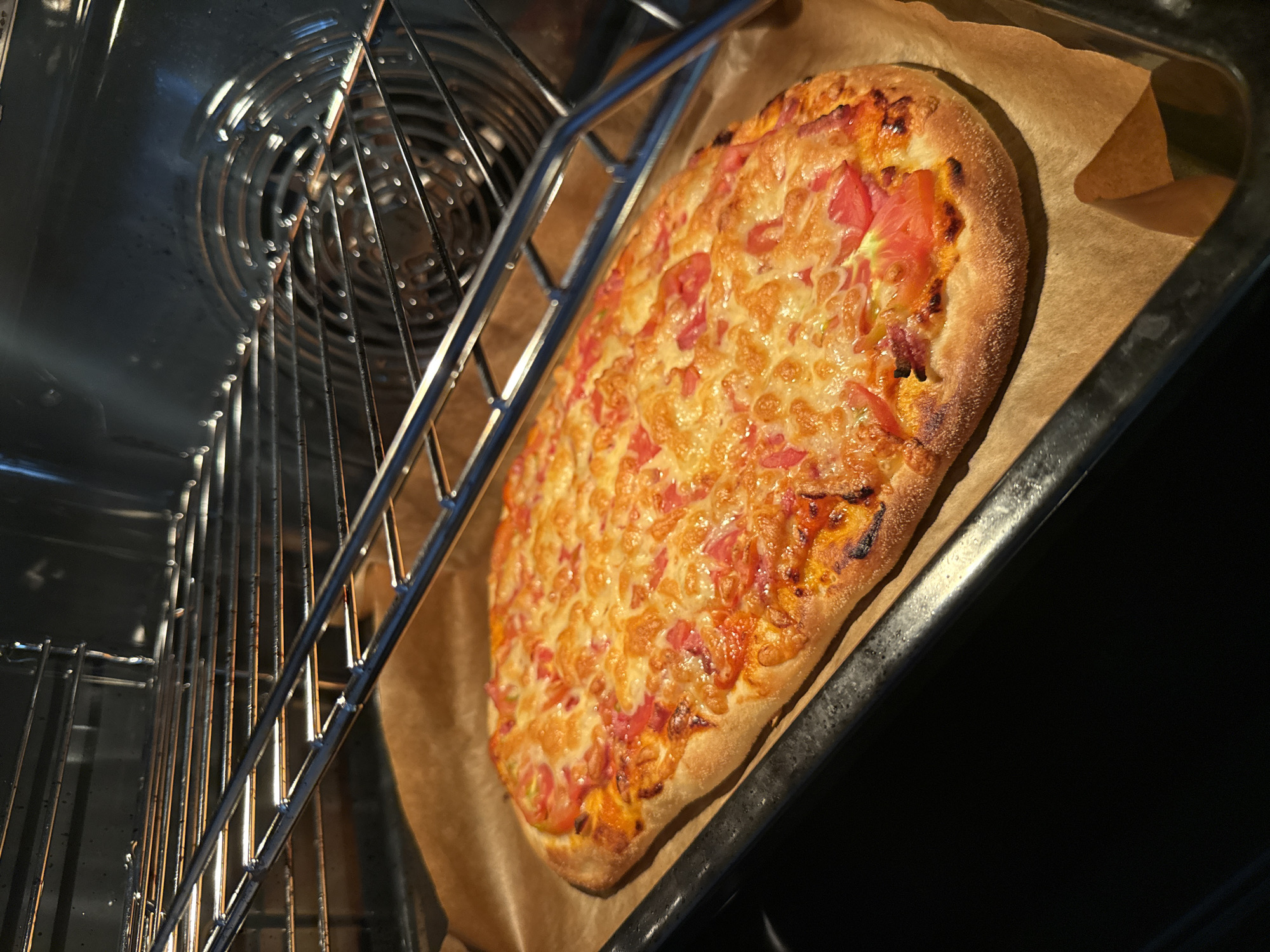 An oval-shaped pizza with melted cheese and tomato pieces is on a baking sheet lined with parchment paper. The baking sheet is inside a dark oven, with oven racks visible above the pizza. The oven interior is metallic and has a fan at the back. The pizza appears to be freshly baked or in the process of baking, with some browned crust edges. The lighting suggests the oven light is on. This scene takes place in a kitchen, likely in Kazan, Russia.