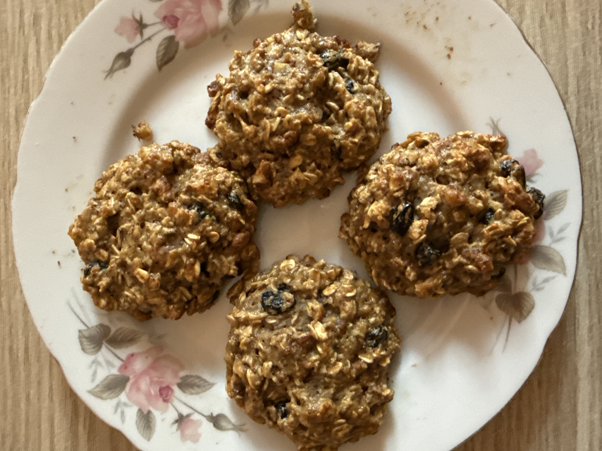 Four oatmeal cookies with visible raisins are arranged on a white plate with a floral pattern. The plate is situated on a wooden surface. The image is a top-down shot. The location is Dilijan, Armenia.