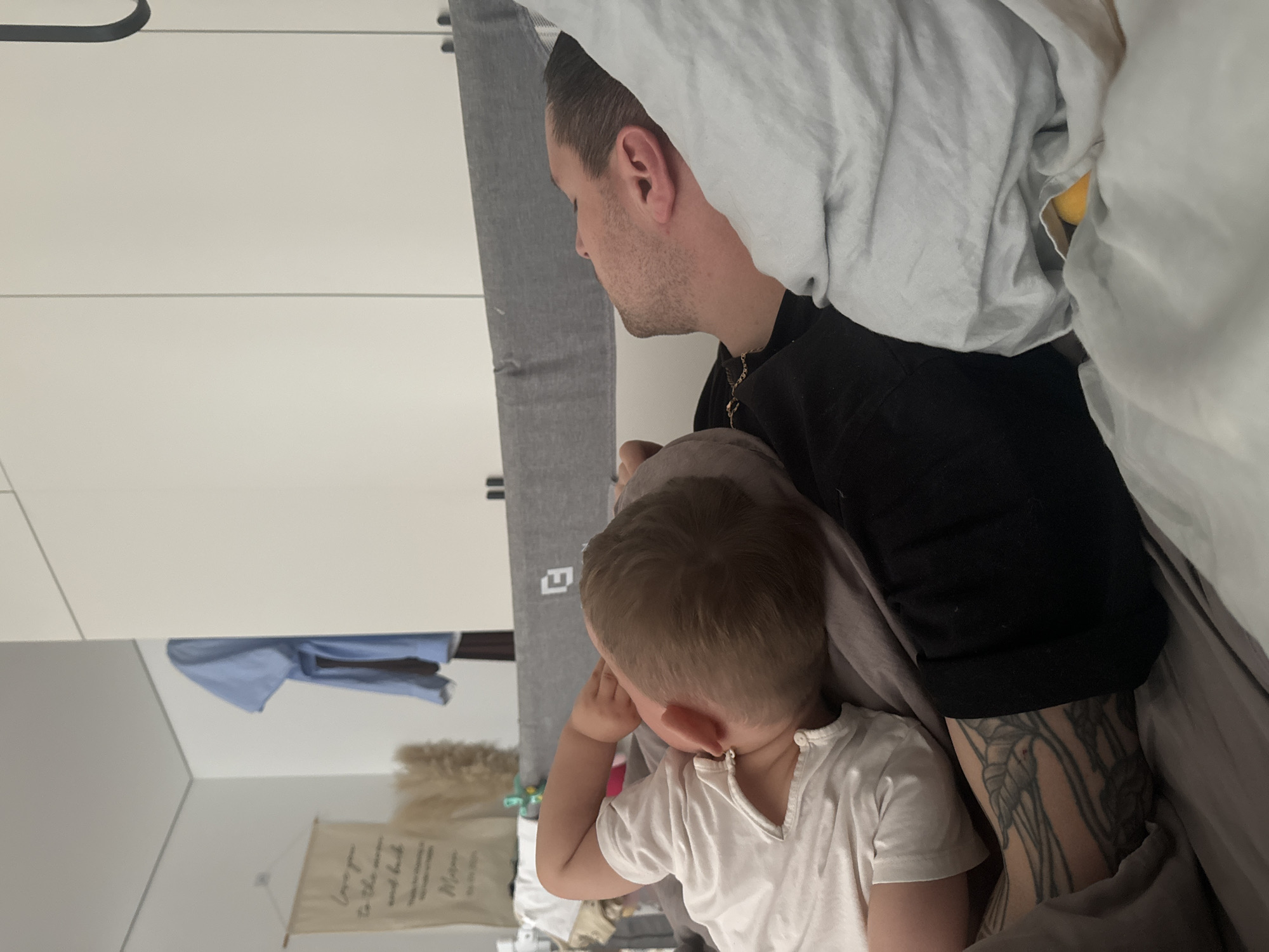 A photograph captures a man and a child lying in a bed. The man, seen in profile, wears a black top and has a tattoo on his arm. A gold chain is visible around his neck. The child, wearing a white shirt, is positioned close to the man. They are covered by grey and white sheets. A wardrobe is visible in the background. A banner hangs on the wall.