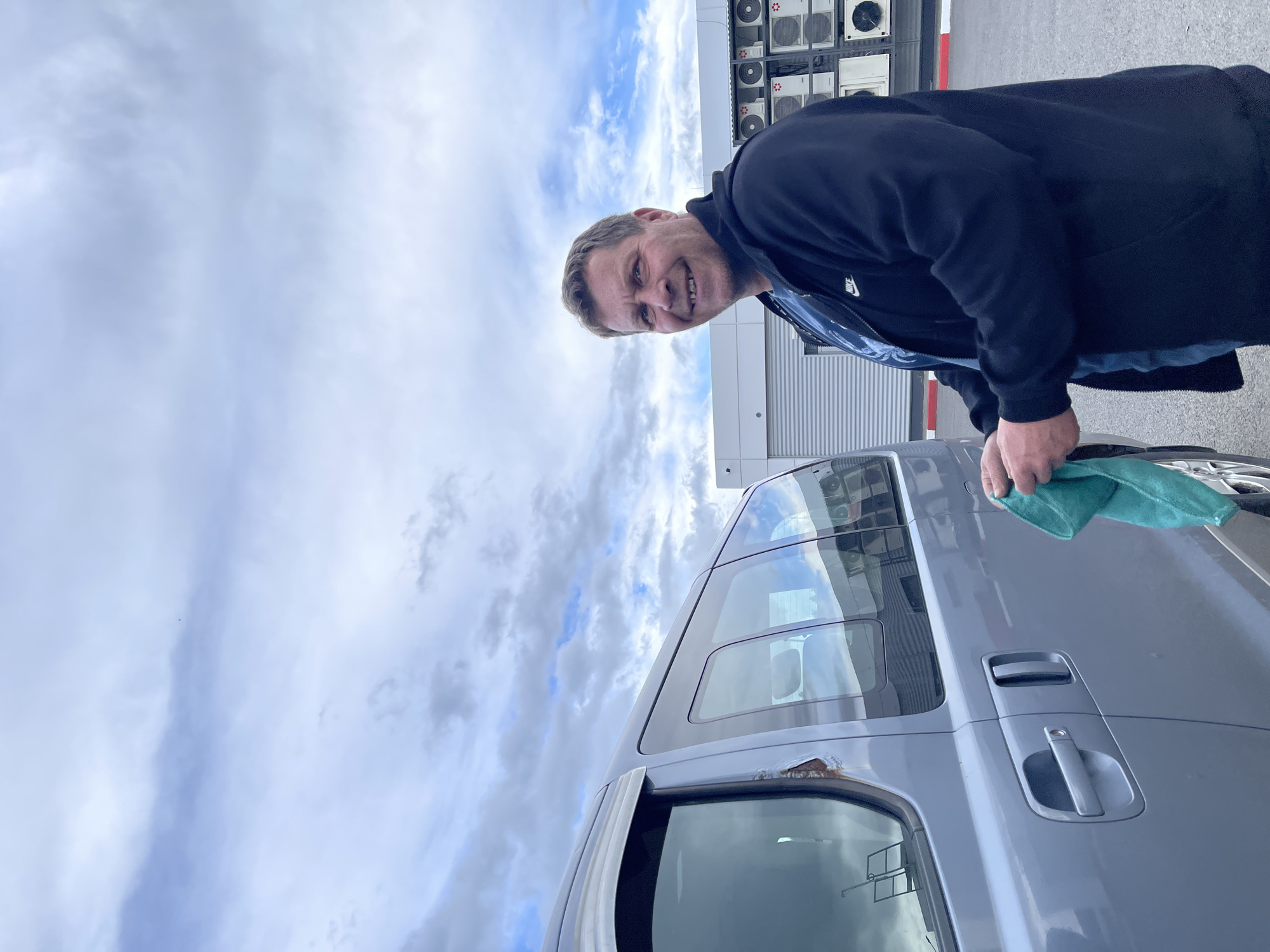 A man is polishing a silver van in an outdoor urban setting. The sky is cloudy, with white and blue hues. The man is wearing a dark jacket. Multiple air conditioning units are visible in the background. The location is identified as Бавыкино, Russia.