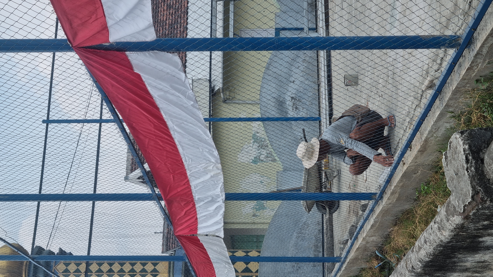 An exterior, eye-level, medium shot shows a person squatting down behind a chain-link fence decorated with a red and white Indonesian flag. The person, wearing a wide-brimmed straw hat, a gray long-sleeved shirt, and dark shorts, is facing away from the camera and appears to be examining or manipulating something on the ground near the fence. A brown bag is slung over their left shoulder. The chain-link fence has blue metal posts and a blue metal railing along the bottom. Beyond the fence, there are what appear to be large, gray tarp-covered structures and a yellow building with a red-tiled roof. In the background, on the left, a portion of a building with a distinctive gold and black geometric pattern is visible. The ground inside the fenced area is paved concrete. The area outside the fence, in the foreground, shows a concrete channel with some sparse grass and dirt. The scene is consistent with an outdoor location in Diwek, Indonesia, possibly a public space or a construction site, given the presence of the tarped structures and the general atmosphere.