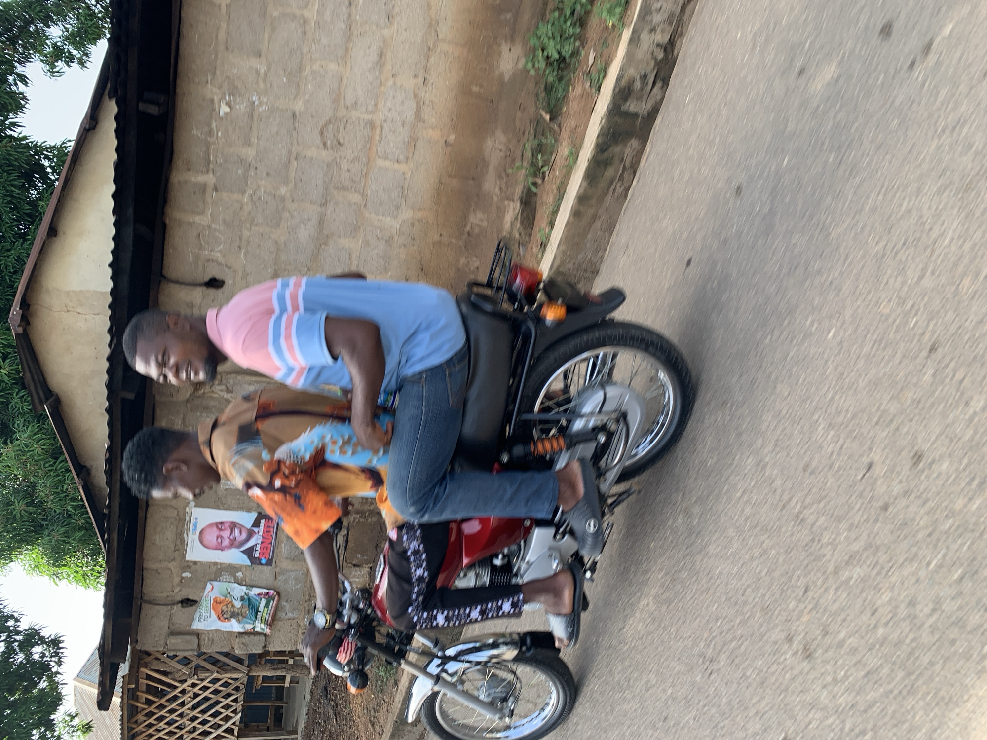Two men are riding a red motorcycle on a paved road. The man in the front wears an orange patterned shirt and black pants with a white pattern. The man in the back wears a blue shirt with pink stripes and jeans. Both men wear sandals. A building with posters of political figures is in the background. The location is Akwanga, Nigeria.
