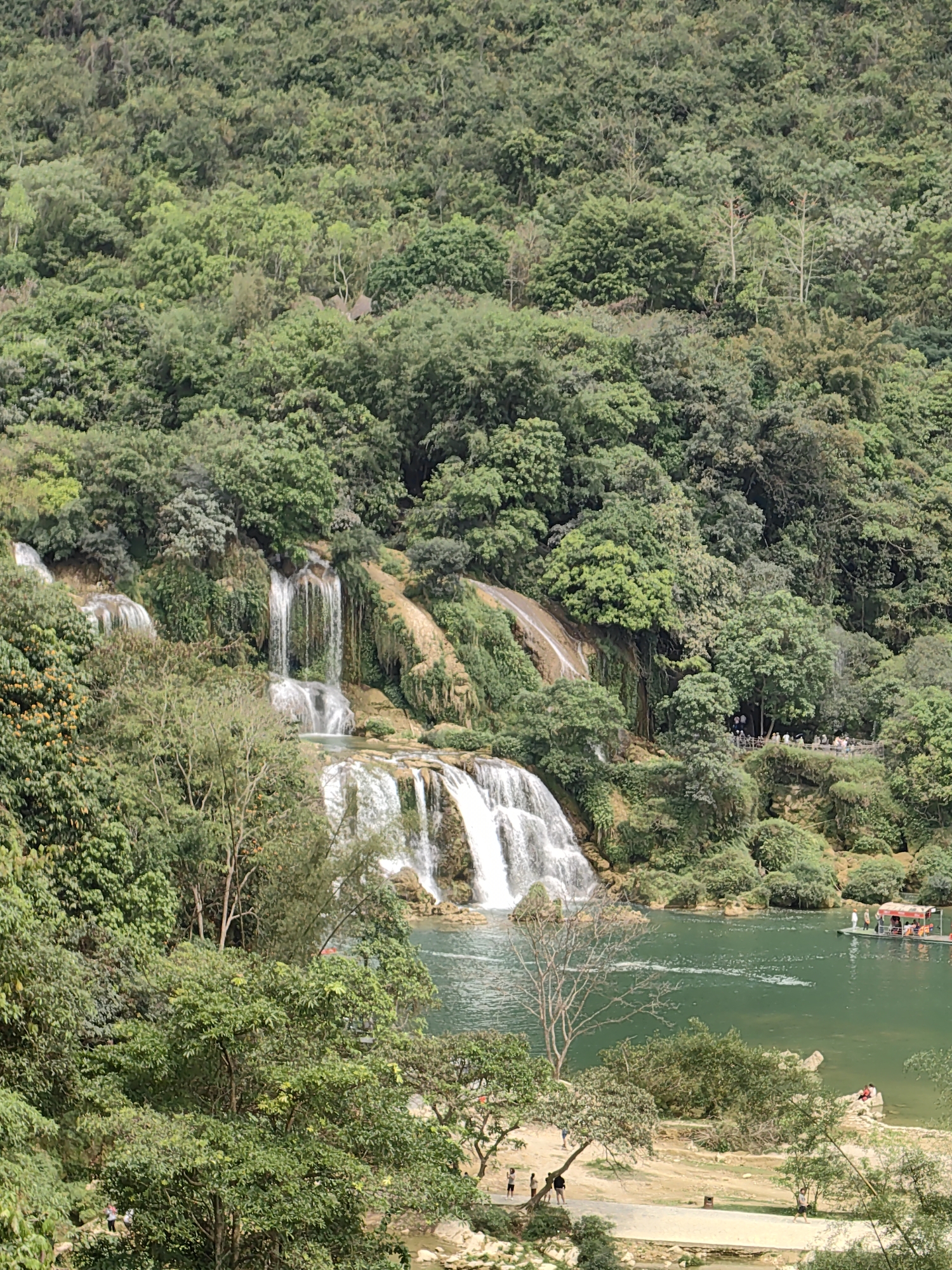 The image shows the Ban Gioc Waterfall in Xã Đàm Thủy, Vietnam. Lush green vegetation covers the hillsides surrounding the waterfall, which cascades down multiple tiers into a green-hued lake. Groups of people are visible along the shore and on a small boat in the lake. A sandy area is visible in the foreground with people walking around.