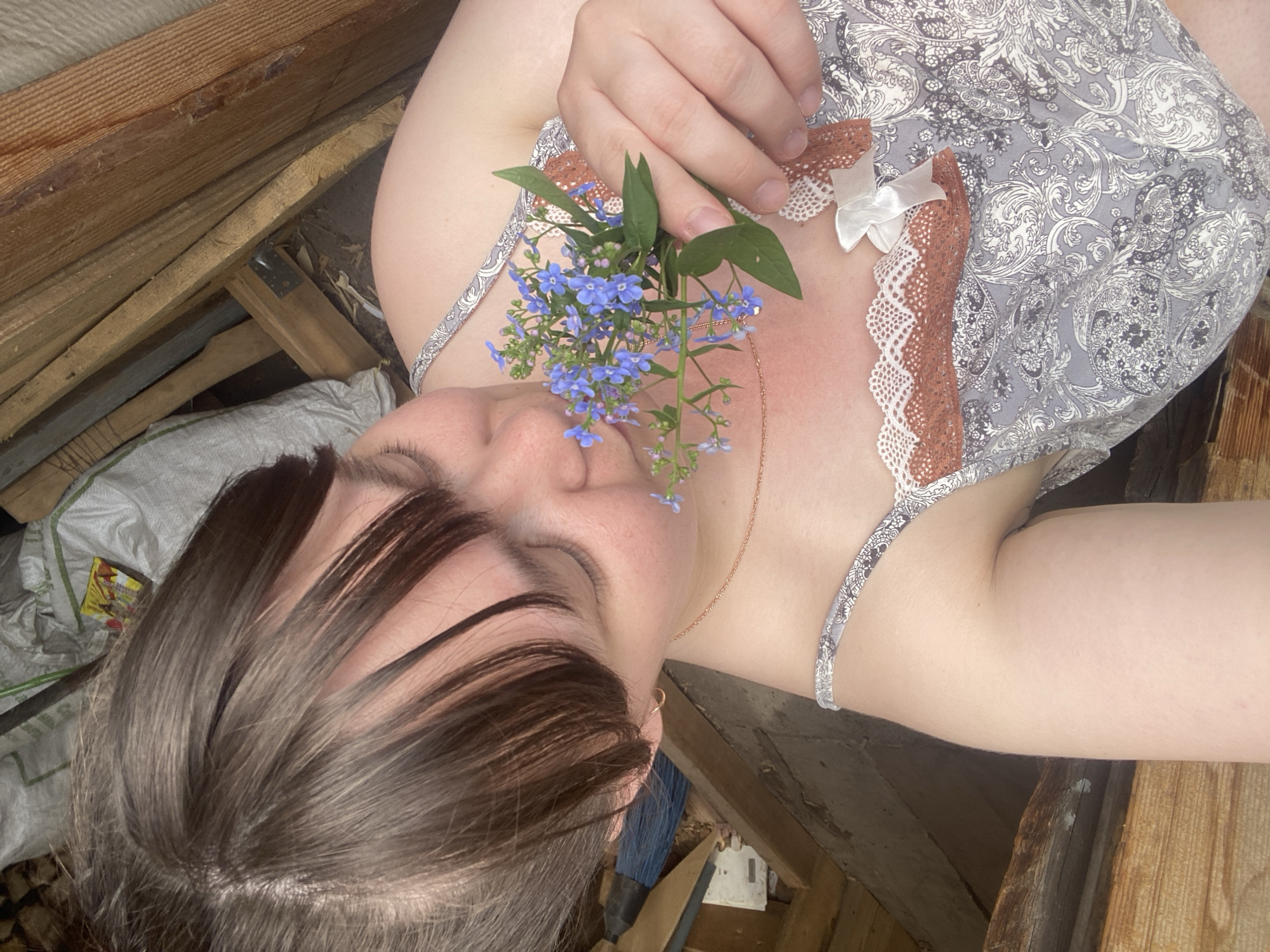 A woman lies sideways in what appears to be a rustic or unfinished attic space. She holds a small bouquet of blue forget-me-not flowers near her face. The woman has dark hair that partially covers her face, and she is wearing a light-colored patterned top with lace detailing. The surrounding environment consists of wooden planks, possibly part of the attic's structure. A white bag and debris are visible in the background. The location is stated as the city of Votkinsk, Russia.