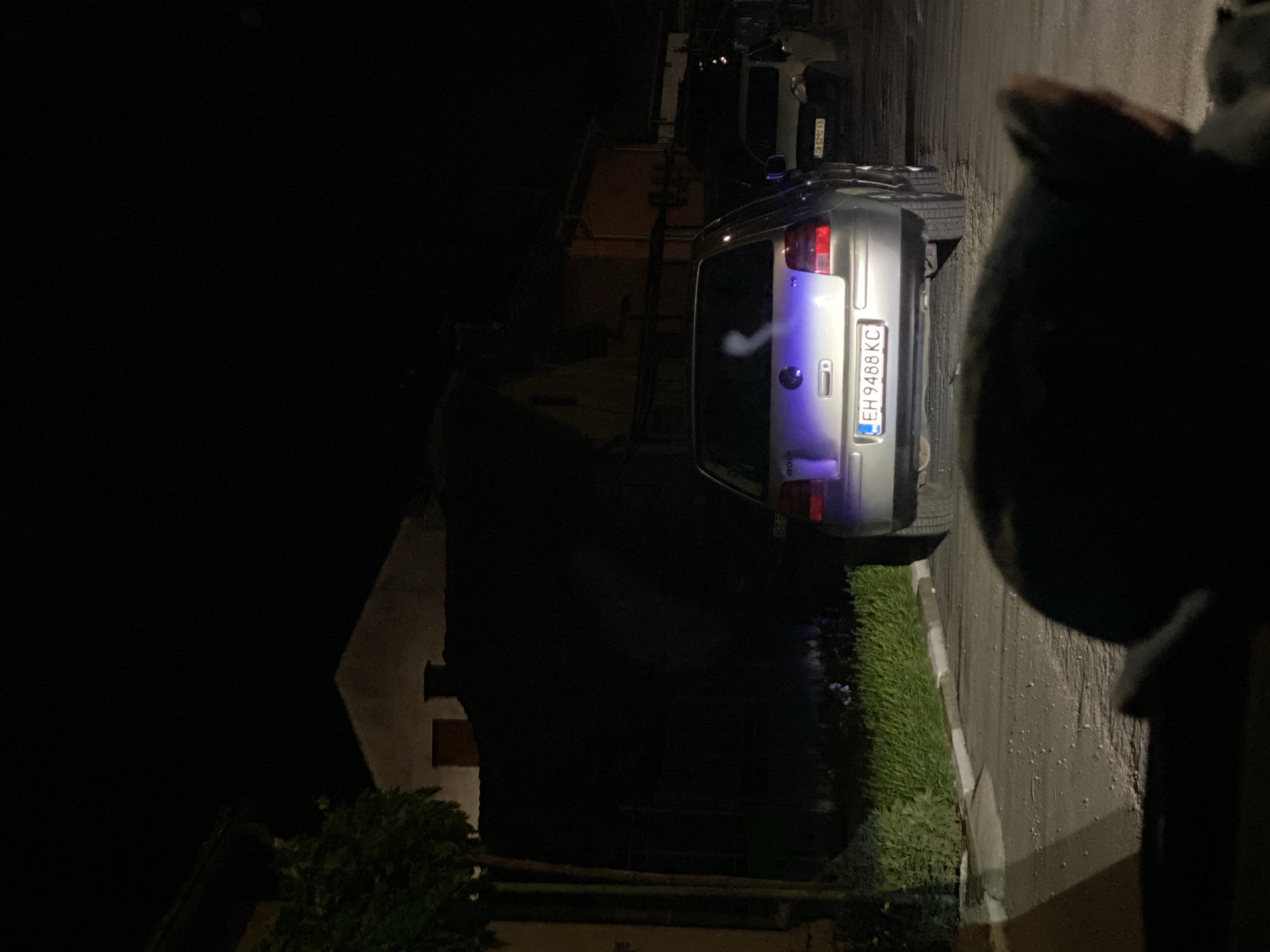 The image depicts a street scene at night in the city of Yasen, Bulgaria. In the foreground, a dark, out-of-focus object, possibly an animal's head or a piece of furniture, is visible. The middle ground is dominated by the rear of a silver Volkswagen Golf car, its license plate reading "EH 9488 KC". The car's taillights are illuminated, and a wispy white cloud, possibly exhaust or steam, is visible above its rear. To the right of the Golf, a lighter colored car is parked. The background shows the dark facades of buildings with dimly lit windows. A green hedge runs along the side of the road in front of the buildings. The street is paved and appears to be sparsely lit, with some scattered white debris on the surface. The overall atmosphere is nocturnal and somewhat muted due to the low light conditions.