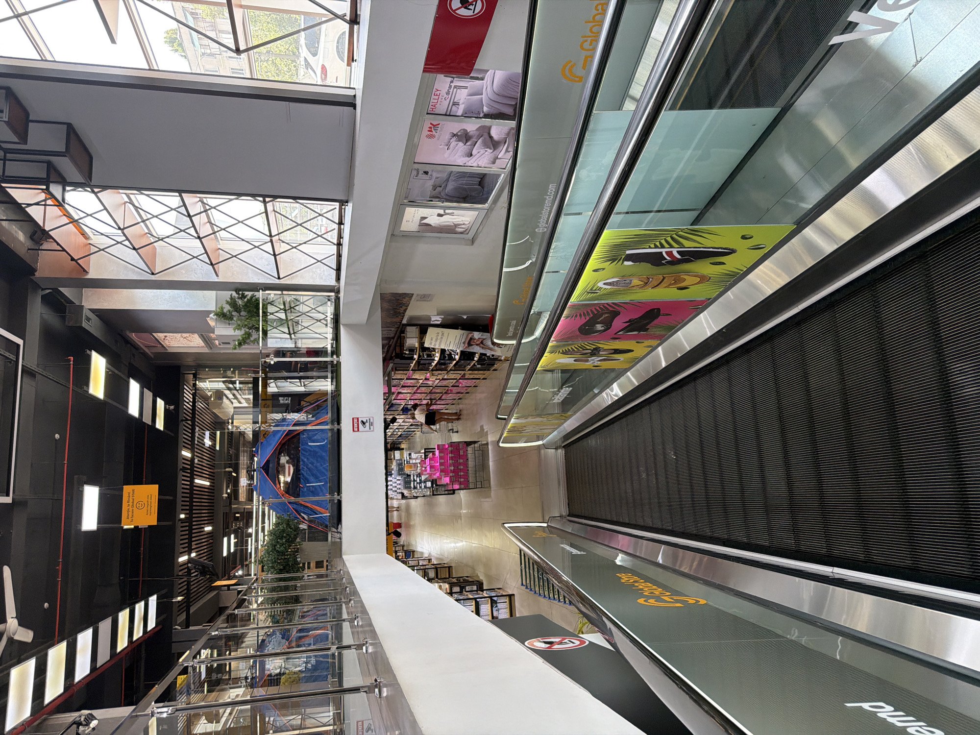 The image shows the interior of a large store, possibly a department store or a hypermarket, with multiple levels. In the foreground, there is a downward-moving escalator with a metallic railing and a glass barrier. Advertisements featuring products like bedding are visible on the glass, and also on the side of the escalator, there are images of shoes and plants.
The upper level of the store is visible beyond the escalator. There are shelves filled with merchandise, and a blue and orange tent is displayed prominently. The ceiling is black with numerous lights. A sign in an orange square with smiling emojis and text in Romanian can be seen hanging from the ceiling, reading "Atenție, toți funcționează!" which translates to "Attention, everything is working!".
In the background, there are aisles of products, and a few people are seen browsing. The architecture features a modern design with glass railings and exposed structural elements. The overall impression is that of a busy retail environment. The context provided indicates the location is in Chișinău, Moldova.