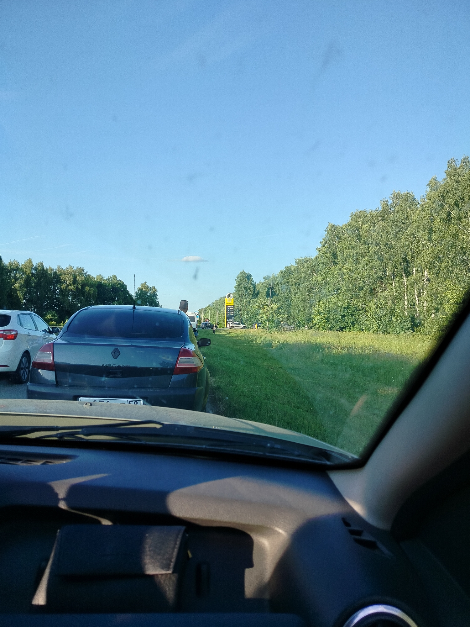 The image shows a traffic jam on a sunny day. The camera is positioned inside a car, looking out through the windshield. In the foreground, the dashboard and part of the steering wheel are visible. In the middle ground, a dark grey sedan is stopped in traffic, with its rear end facing the camera. The license plate is partially obscured but appears to have Russian characters. Other cars are visible further ahead in the traffic jam. To the right of the road, a dense line of trees forms a forest. A bright yellow sign is visible in the distance on the right side of the road, possibly indicating a gas station or a service area. The sky is clear blue with a few wispy clouds. The overall scene depicts a common occurrence of road congestion in a rural or semi-rural setting, possibly near Lunino, Russia, given the presence of Russian license plates.