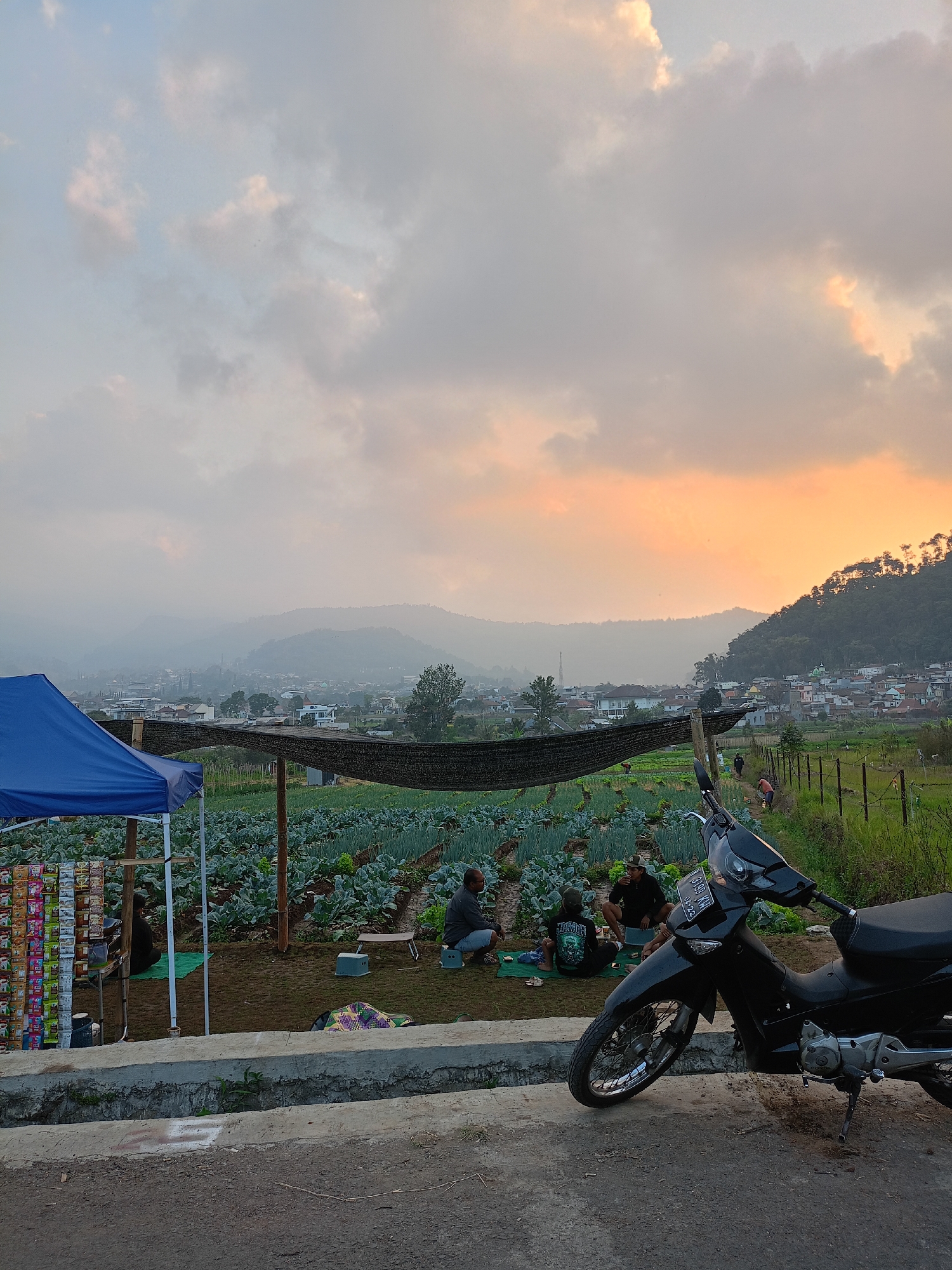 A rural scene in Batu, Indonesia, captures a moment at sunset with a dramatic sky of orange and grey clouds. In the foreground, a black motorcycle is parked on a concrete surface. To the left, a blue tent is set up, displaying various packaged goods on shelves. Several individuals are seated on the ground near the tent, some on mats, and one person is seated on a small stool. Beyond this area lies a field of crops, likely vegetables, with a dark net covering a section. In the mid-ground, a hazy view of a town nestled among rolling hills and mountains is visible. The overall atmosphere is tranquil, with the fading light casting a soft glow over the landscape.