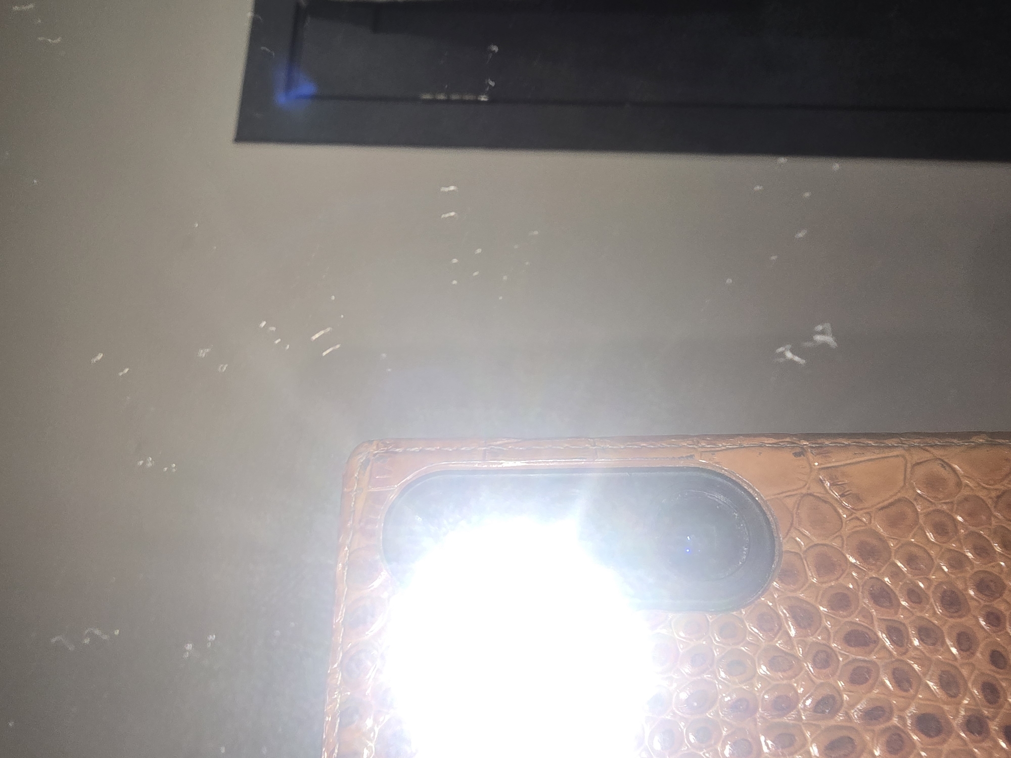 The image shows a close-up of a light brown, textured phone case with a bright light emanating from the camera lens area. The case has a crocodile-like pattern. The background is a light gray wall with several small, scattered white specks or marks. To the right, a dark vertical object, possibly a door frame or edge of furniture, is visible. The lighting suggests the photo was taken with the phone's flash on. There are no people or explicit geographical markers visible that would allow for identification of the location as Ulan-Ude, Russia.