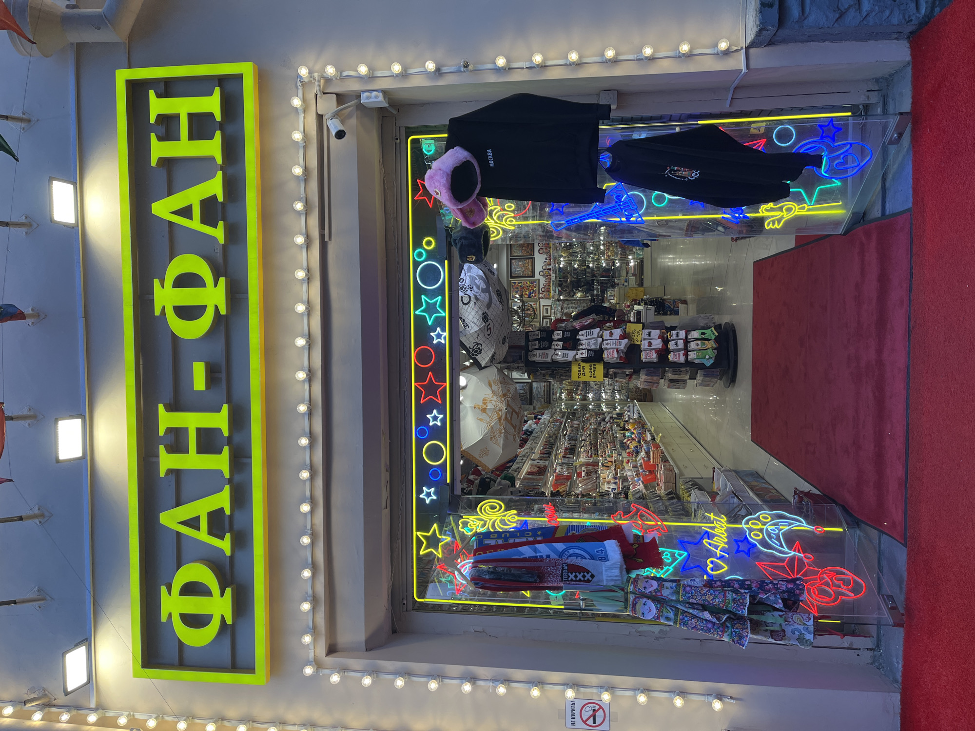 Here is a description of the image:
The image shows the storefront of a souvenir shop named "ФАН-ФАН" (FAN-FAN), located in Moscow, Russia. The shop's name is displayed vertically on a bright yellow sign with yellow neon lights. The storefront is decorated with neon signs in various shapes, including stars, circles, and cartoon figures. There are also items for sale displayed in the shop window, such as hats, scarves, and small souvenir items. A red carpet leads into the shop. A surveillance camera is mounted above the entrance. A no smoking sign is posted in the lower left corner of the window.