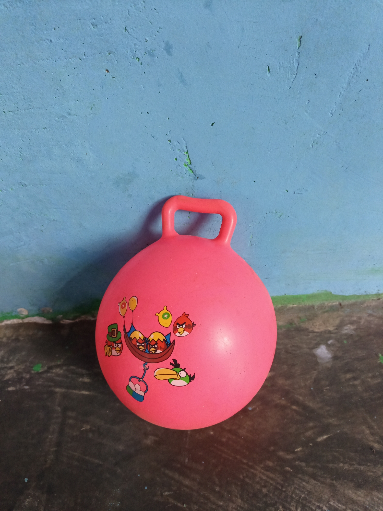 A bright pink, inflatable ball with a handle sits on a dark, textured surface in front of a blue wall. The ball is decorated with cartoon images of Angry Birds characters, including several birds in a basket and a bird with a yellow beak and green wings. The blue wall appears textured and slightly weathered, with some green paint streaks visible near the bottom. The dark surface beneath the ball has a rough, almost concrete-like texture, with some lighter specks and debris. The scene is lit from above, casting a slight shadow from the ball onto the surface. The overall setting suggests an outdoor or partially covered area, possibly a courtyard or patio. There is no indication of the specific location within Terbanggi Marga, Indonesia, other than the general visual cues of the objects and environment.