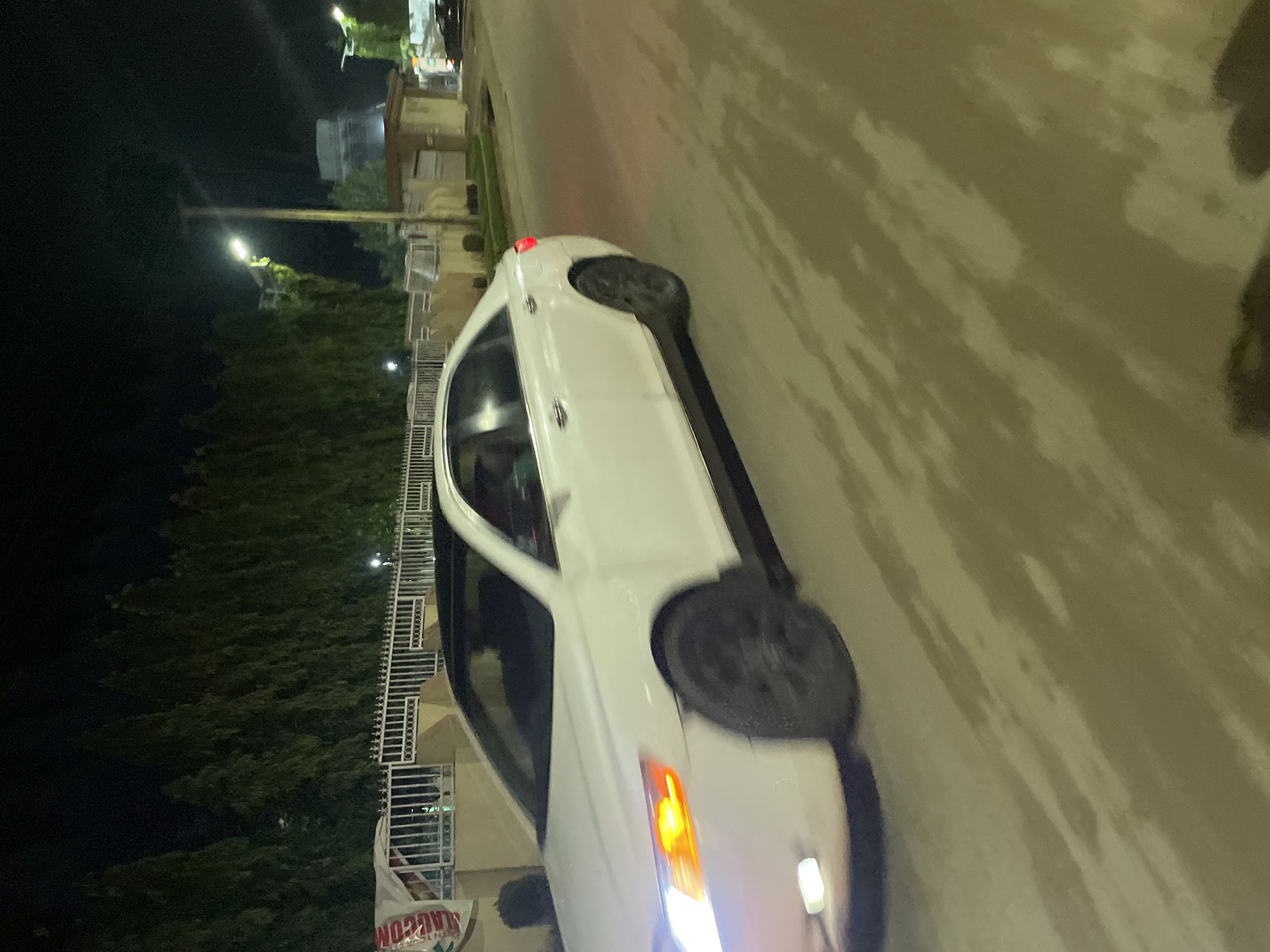A nighttime scene in Abuja, Nigeria, features a white sedan parked or slowly moving on the right side of an asphalt road. The car's bright white headlights and orange indicator light are illuminated at the front, while a red taillight is visible at the rear. The road surface reflects light, showing patterns of illumination likely from streetlights.
To the left of the car, across the road, a line of lush green trees stretches into the dark background. Beyond the trees and further into the distance, several buildings are faintly visible under the dim night sky, some appearing to be residential structures. Bright streetlights punctuate the darkness, casting pools of light onto the foliage and structures.
Adjacent to the road and in front of the trees on the left, a white metal fence or gate runs parallel to the street. Attached to this fence is a partially visible white banner. The text on the banner reads "MAUCON" and "GREEN YES," although parts are obscured and the text is slightly inverted due to the original image's orientation. The overall ambiance is quiet and illuminated by artificial light sources. No people are visible.