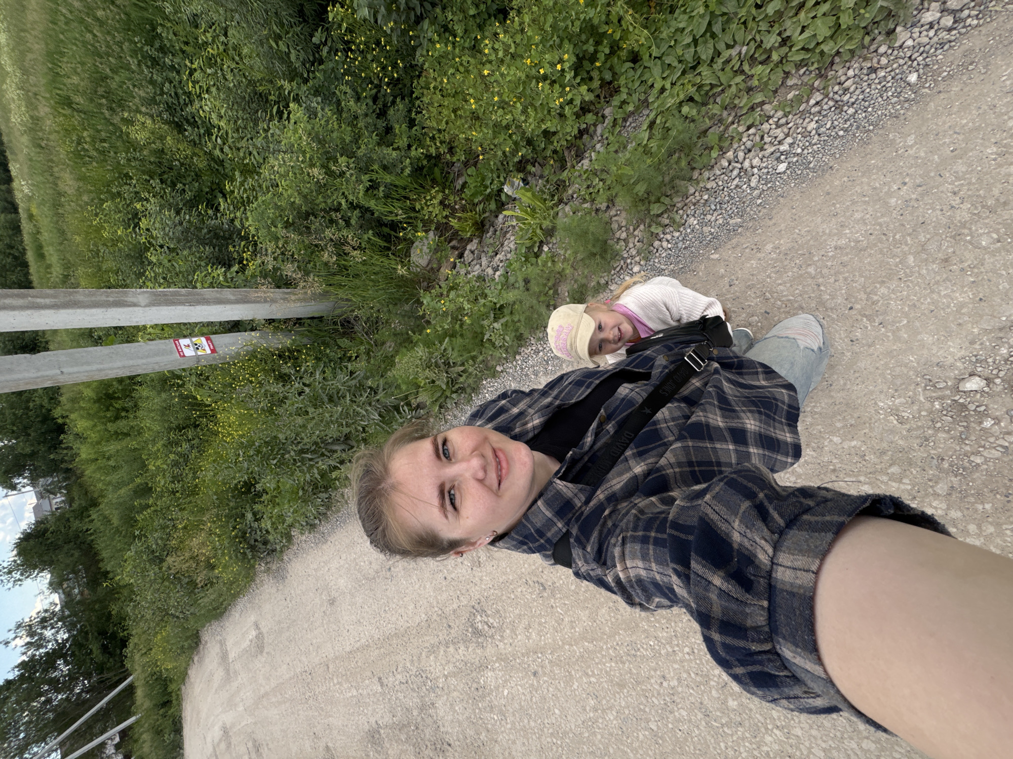 In a selfie taken from a slightly high angle, a woman with blonde hair pulled back and blue eyes smiles at the camera. She is wearing a dark plaid shirt over a black top and light blue jeans. A black bag with a strap displaying "DAVID JONES" is slung across her chest. To her right, a young girl with blonde hair and a light pink hat smiles, wearing a white knit sweater over a pink shirt. They are standing on a light-colored gravel road. To the right of the road, dense green vegetation with small yellow flowers grows, and two concrete utility poles stand among the greenery. One pole has a red and white warning sign attached. The background shows more green foliage and trees, with glimpses of buildings in the distance under a cloudy sky. The overall scene appears to be a rural or suburban road in or near the city of Gorniy, Russia.