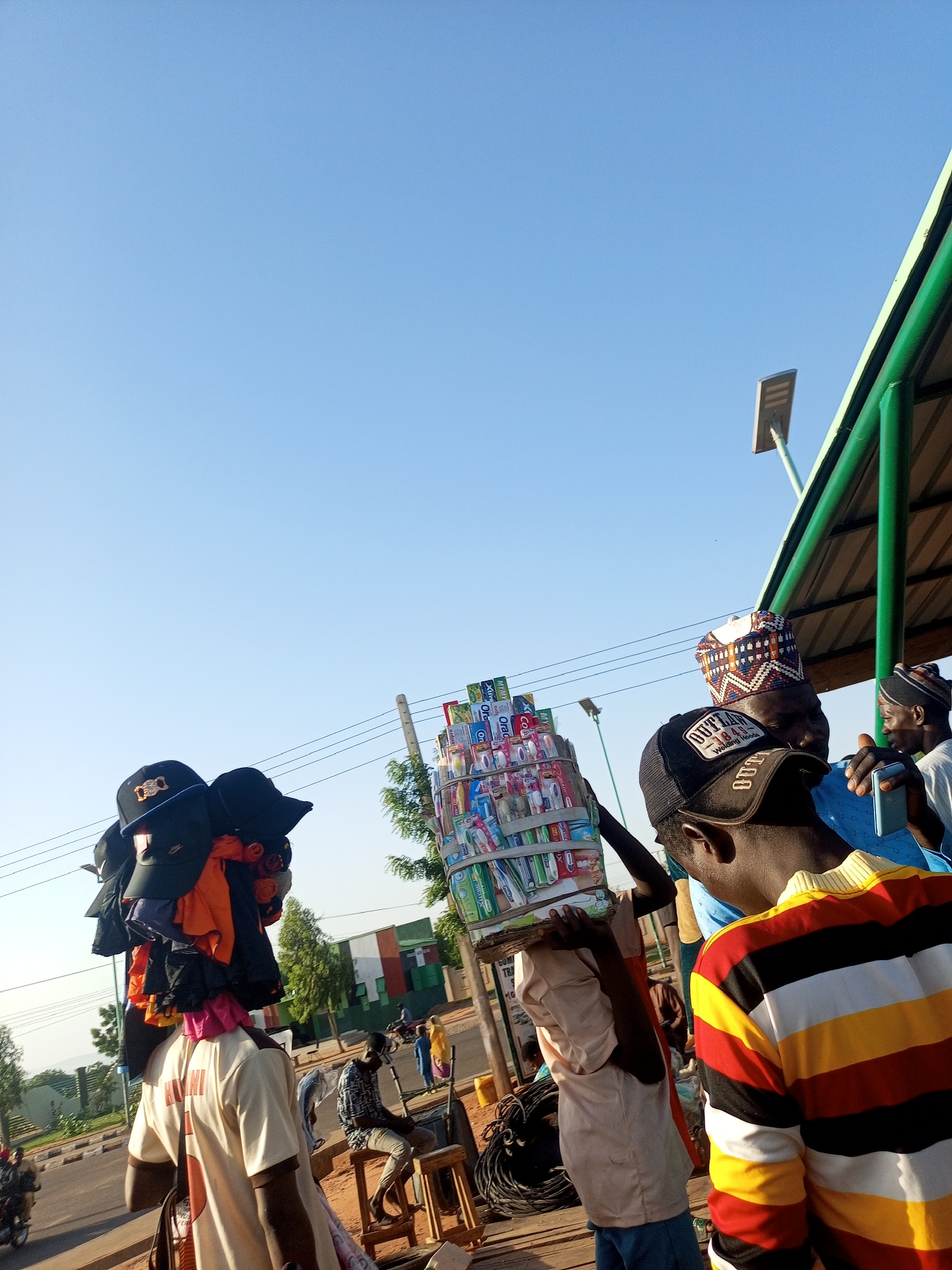 The image captures an outdoor scene in an unknown city. A man is carrying a large stack of toothbrushes on his head. To the left, another man balances a pile of hats and clothing. Several people are gathered nearby, including one in a striped shirt. A seated individual and other pedestrians are visible in the background. The environment includes a paved area with street infrastructure and trees.
