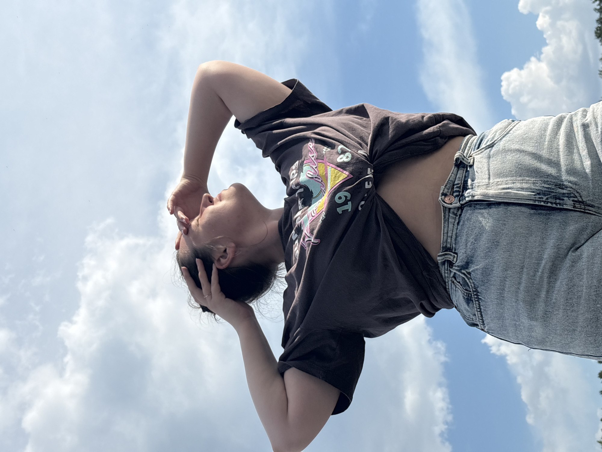 An outdoor, low-angle, medium shot shows a young woman from the waist up, against a bright blue sky with scattered white clouds. She is looking upwards and to the right, with her right arm bent and her hand shielding her eyes from the sun. Her left hand is in her dark hair, which is pulled back. She is wearing a dark, short-sleeved t-shirt tied at the waist, exposing her midriff, and light-wash denim shorts. The t-shirt has a graphic print on the front, featuring text and colorful abstract shapes. Her expression is serene as she enjoys the daylight. The image was captured in Chaykovsky, Russia.