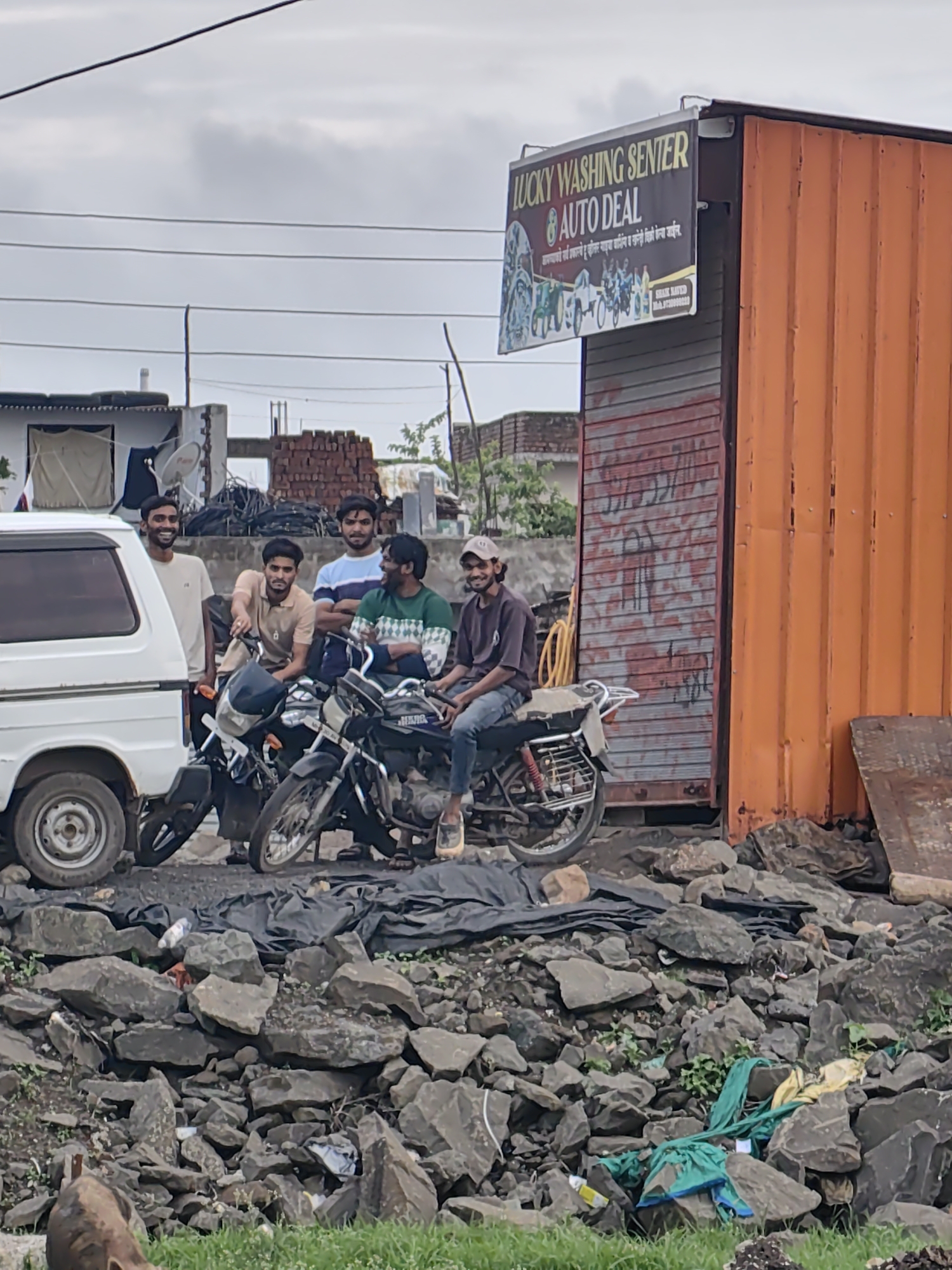 Four young men are gathered around two motorcycles in front of a storefront. The storefront is an orange corrugated metal shipping container with a sign that reads "LUCKY WASHING CENTER AUTO DEAL". The men are casually dressed, and one is sitting on a motorcycle while the others stand or lean against the bikes. They appear to be interacting and possibly waiting. A white van is parked to the left of the motorcycles. The ground is covered in rocks and debris, with some green fabric scraps visible in the foreground. The sky is overcast. The location is identified as Walsawangi, India.