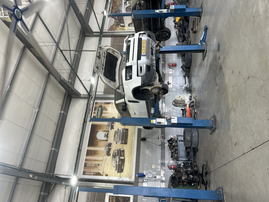 An auto repair shop features a white Land Rover Defender elevated on a blue lift. Its hood is open, revealing the engine bay. Car parts and tires are scattered around the shop floor. A poster showcasing various Land Rover models adorns the wall. Another car is being worked on nearby. The shop's interior includes a metal roof structure and bright lighting.