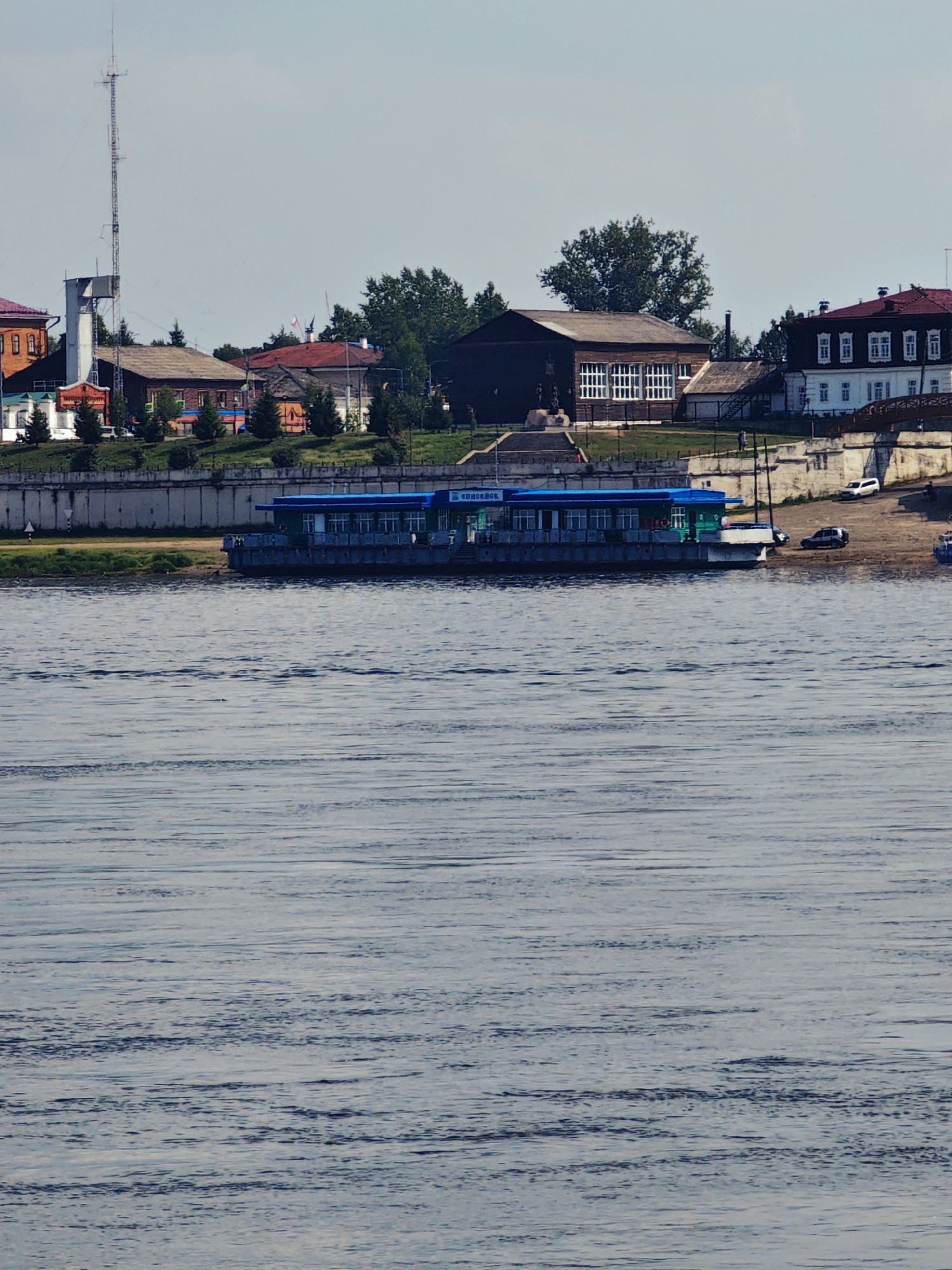 The image shows a wide, calm river in the foreground. In the middle ground, a green and blue boat or ferry is docked at a concrete embankment. Beyond the embankment, a city skyline is visible, featuring various buildings, trees, and a tall radio tower. The architecture suggests a historical or provincial Russian setting. On the riverbank, there are a few cars parked, and a pedestrian walkway is evident. The sky is partly cloudy. The scene is likely a view of the city of Yeniseysk, Russia, situated on the Yenisei River, a major waterway. The overall impression is one of a tranquil day in a riverside town.