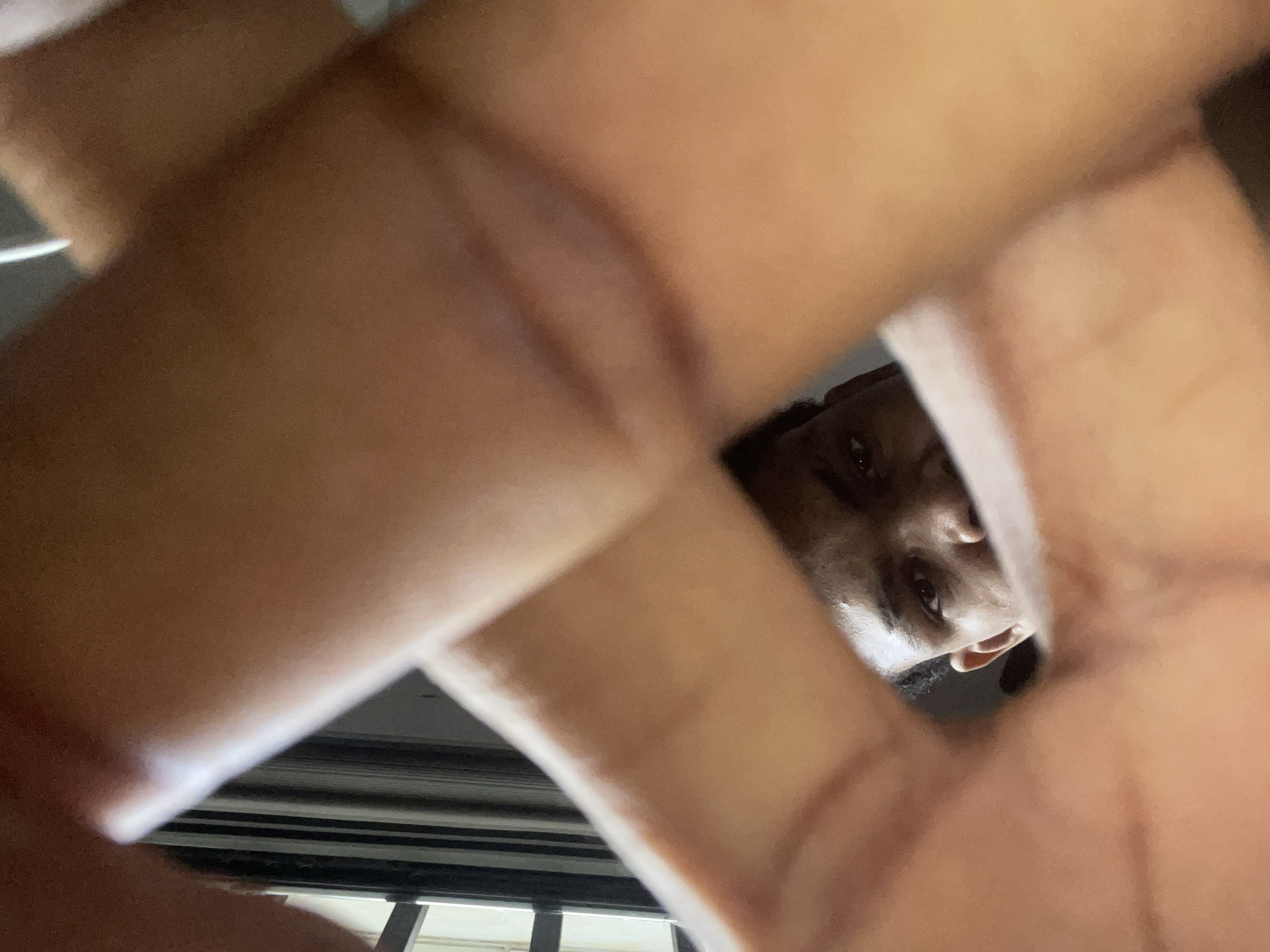 A close-up, low-angle shot of a person's face partially visible through a grid formed by the person's fingers. The person has dark skin and appears to be looking directly at the camera. Behind the fingers and face, a blurry view of what may be a window or barred opening is visible. Given the request to specify location as Ado, Nigeria, the bars could indicate a residential setting. The setting is somewhat dark, with light filtering in from the window or opening.