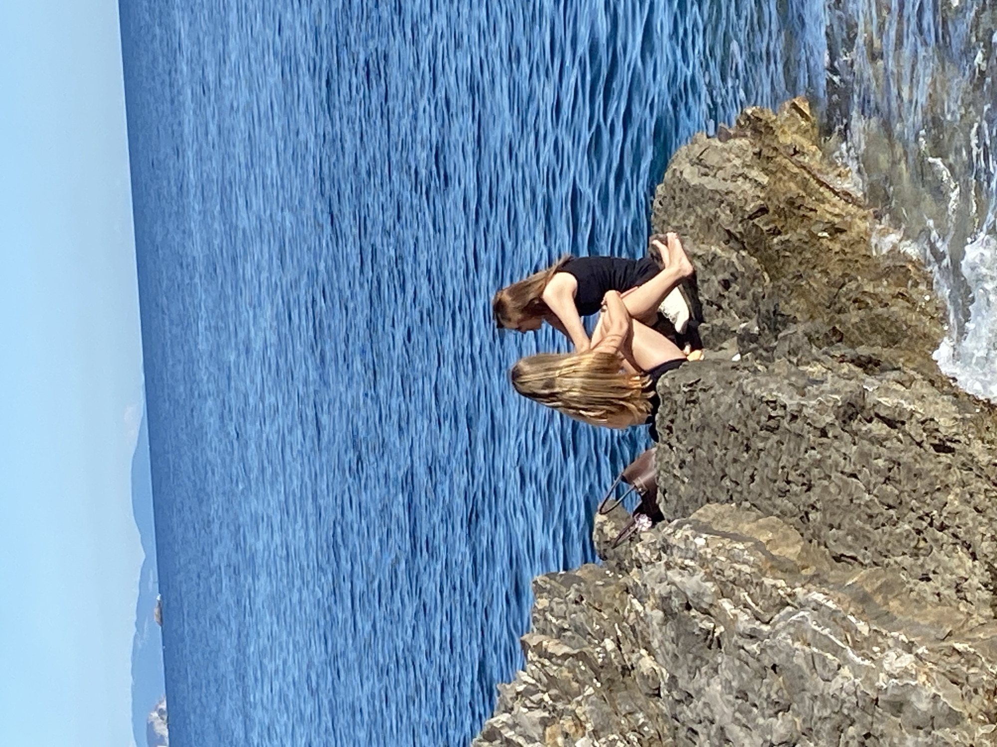 Two people are on a rocky cliff edge next to the sea, potentially in Budva, Montenegro. One person, wearing a black top, is leaning over another person with long blonde hair. The surrounding environment consists of a blue sea and a rocky terrain.