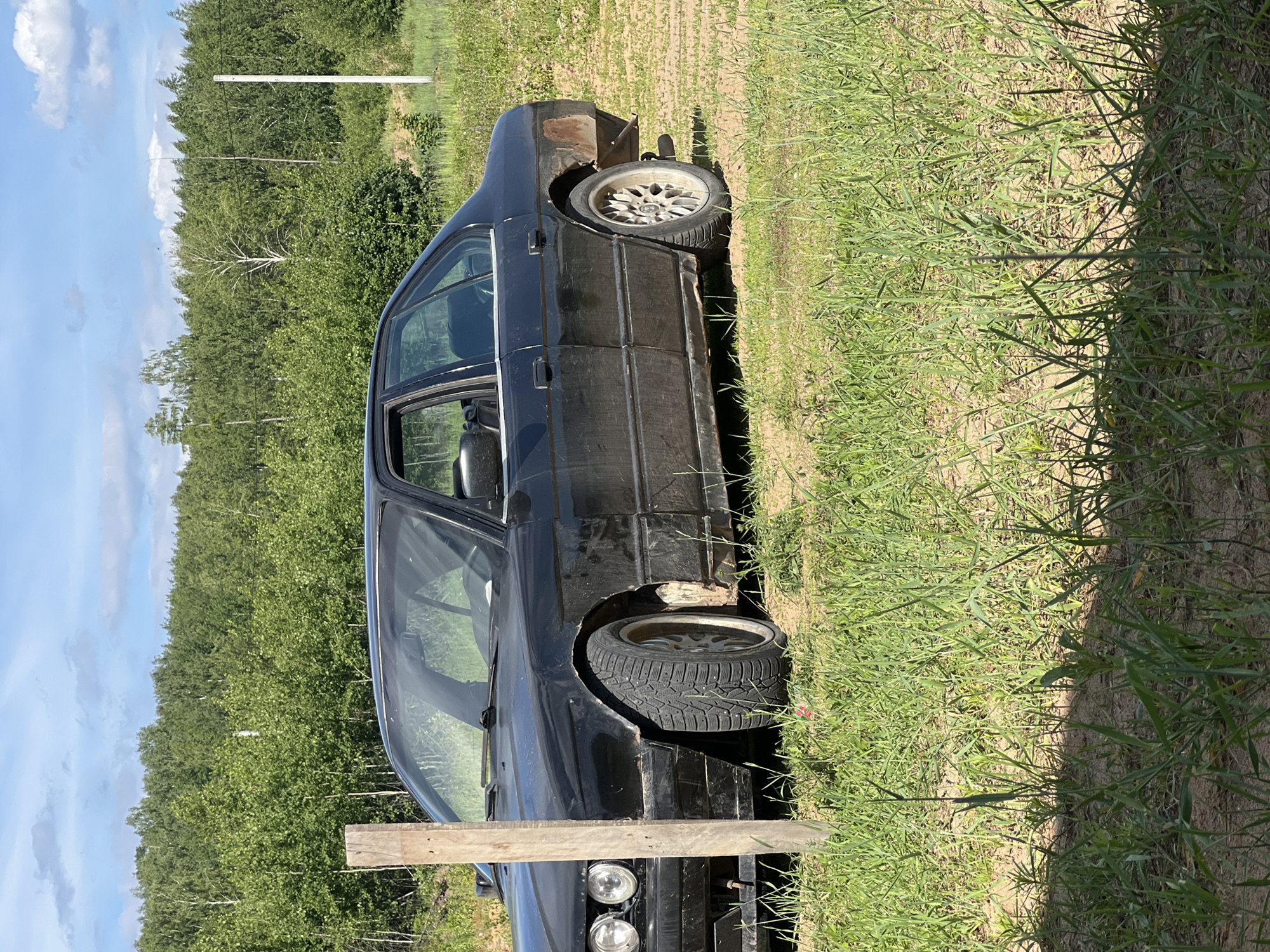 The scene is set outdoors in Noviy, Russia, under a bright blue sky dotted with scattered white cumulus clouds. A black, four-door BMW E34 sedan, in a state of significant disrepair or modification, is parked diagonally on an unpaved area consisting of sparse green grass and light brown soil. The car's front left fender is heavily rusted and appears partially cut away, revealing the wheel. The front left wheel features a multi-spoke alloy rim and a studded winter tire. The vehicle's rear bumper is missing, exposing rusted metal on the rear body panel and a singular exhaust pipe. The driver's side front window is open.
In the immediate left foreground, a weathered wooden post stands vertically, partially obscuring the front end of the sedan. The ground around the car and in the foreground is uneven, showing patches of bare earth mixed with grass. Beyond the car, a dense line of green deciduous trees forms a natural backdrop, suggesting a forest edge. A particularly prominent white-barked tree is visible among the other green foliage in the background. On the far right, a tall, slender white pole extends vertically above the treeline. The scene is illuminated by strong sunlight, casting defined shadows in the foreground. No people are visible.