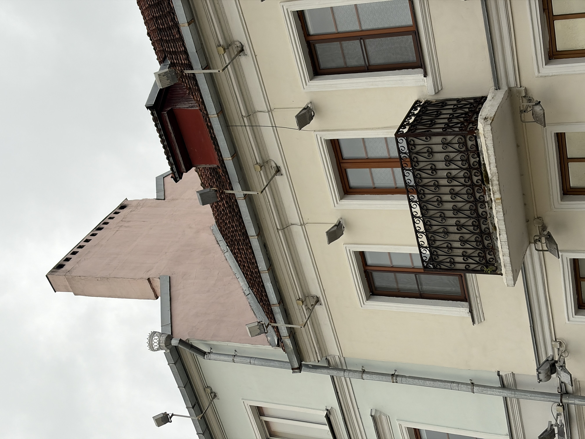 The image shows the facade of two adjacent buildings against an overcast sky. The building on the left is painted a pale green and features a metal downspout running vertically along its exterior. The building on the right is painted a cream color and has a balcony with an ornate black metal railing projecting from its second story. Both buildings have multiple windows with wooden frames. Above the cream-colored building, a section of a red-tiled roof is visible. A pink chimney extends upwards from the pale green building, with a decorative element at its top. Several modern floodlights are mounted on the exterior walls and under the eaves of both buildings. The scene appears to be a street in the city of Minsk, Belarus, based on its architectural style and the details of the buildings.