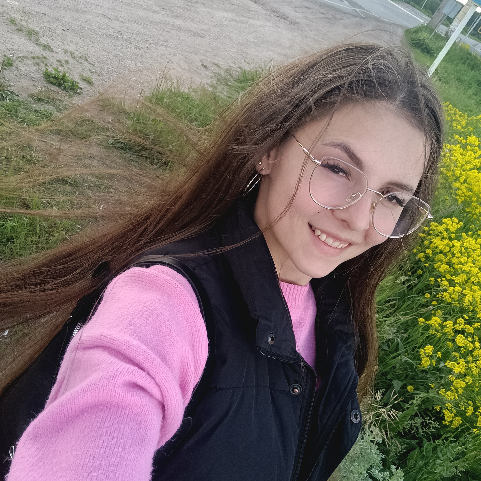 A young woman with long brown hair and glasses smiles into the camera, dressed in a pink sweater and black vest. She is standing outdoors with green grass and yellow flowers to her right. Behind her is a dirt path or road. The location appears to be near a road with sparse vegetation.