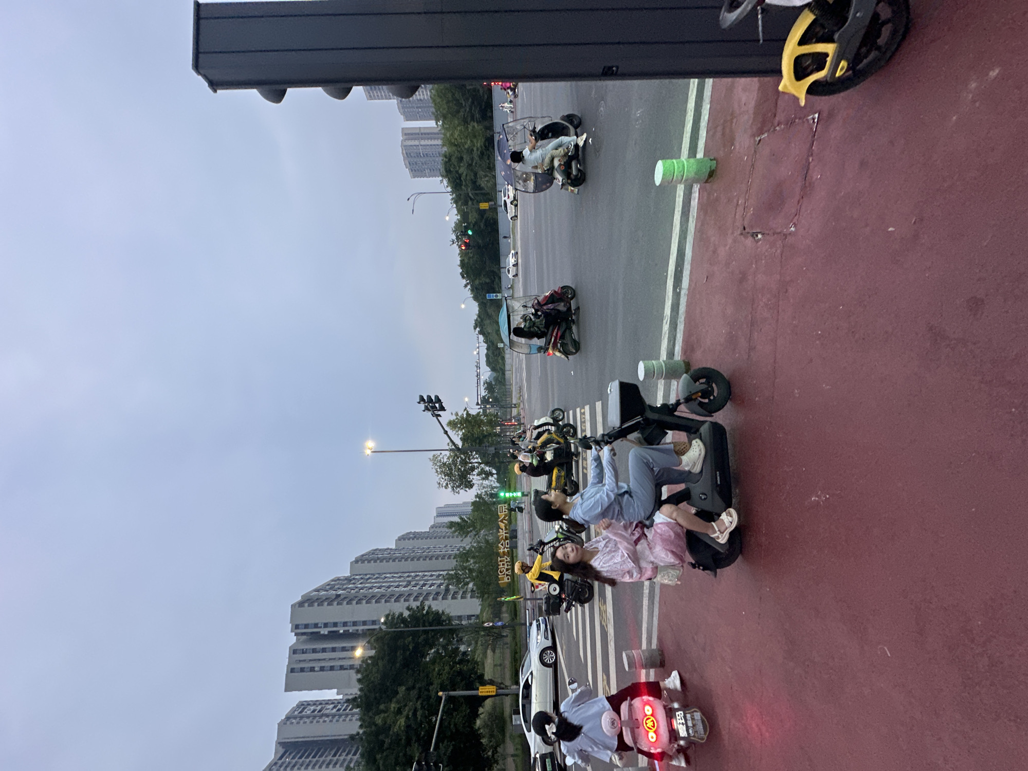 The image captures a street scene in the Chongqing High-tech Zone, China. In the foreground, a red-colored pedestrian path is visible, with several electric scooters and motorcycles on it and on the adjacent road. Two young women are riding a dark-colored electric scooter. The woman in the front is wearing a light blue tracksuit and white crocs, while the woman behind her is wearing a pink dress and white sandals. To their left, another electric scooter is parked, with its red taillights illuminated, and a woman in a light blue top and white pants is seated on it. Further back, a white car is stopped at a crosswalk. Several other electric scooters and a man in a yellow jacket on a motorcycle are seen navigating the intersection. In the background, tall apartment buildings line the cityscape, and streetlights illuminate the scene under a cloudy sky. The overall environment suggests a typical urban setting in China, possibly during the evening or early morning hours.