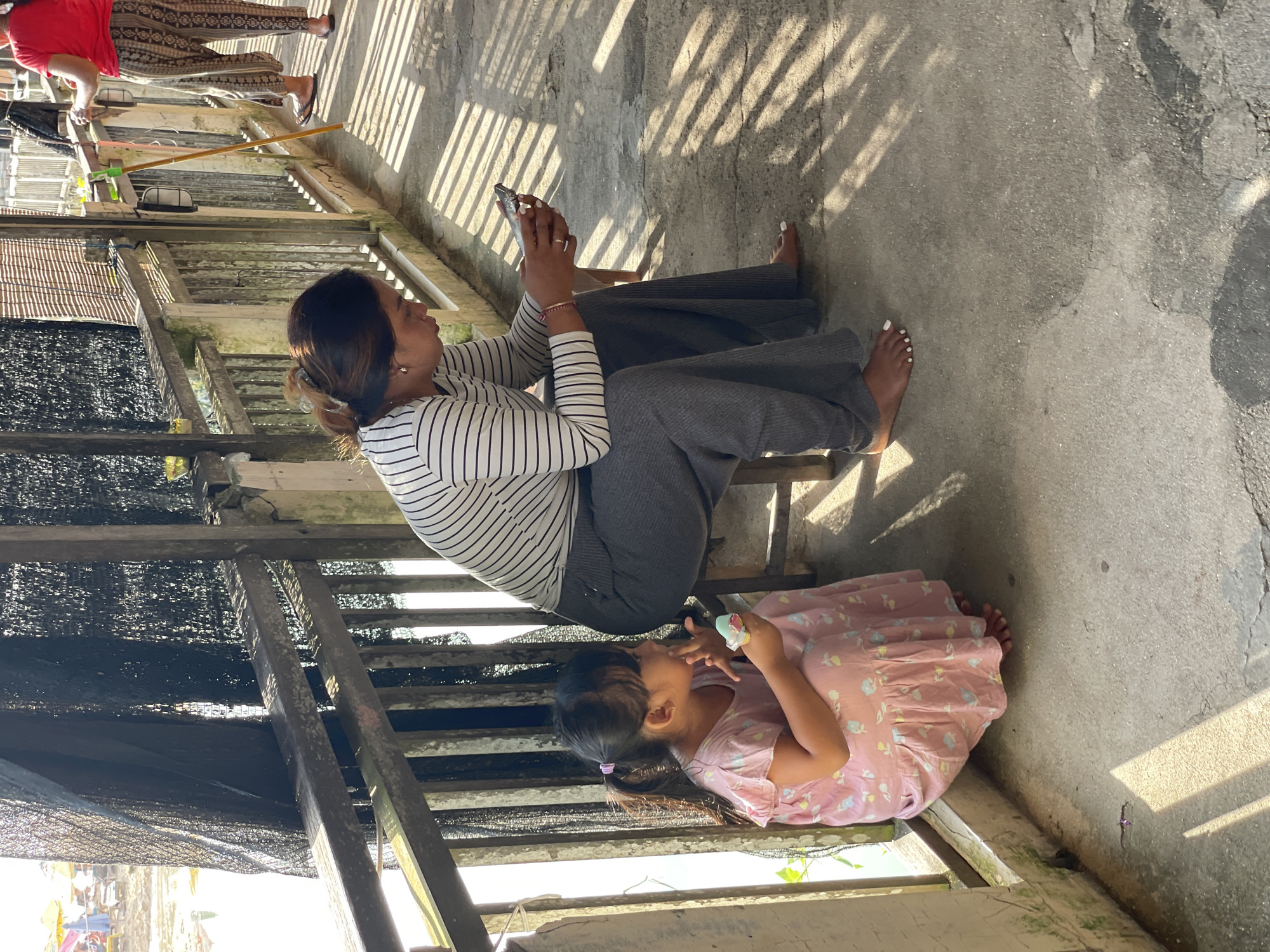 The image depicts an outdoor scene with an adult woman, a young girl, and a partially visible third person under bright sunlight. The main subjects are seated on or next to a wooden structure, possibly a balcony, porch, or elevated walkway.
In the foreground, a woman with brown hair tied back is seated, looking at a smartphone held in both hands. She is wearing a white and black striped long-sleeve shirt and dark grey wide-leg pants. Her feet, with white painted toenails, are visible on the concrete ground. Next to her, a young girl in a pink patterned dress with a ponytail is also seated, holding a small, light blue container or cup and appearing to eat or drink from it.
The setting features a concrete ground or pathway, across which strong, striped shadows from an overhead structure are cast, indicating bright sunlight. A wooden railing and support beams are prominent, with black mesh netting visible underneath, suggesting a protective barrier or shading. In the background, another person is partially visible, standing and wearing a red top and patterned pants, with flip-flops on their feet. The overall environment appears to be a casual outdoor area, possibly within a residential or semi-urban setting, consistent with the Kuta Selatan, Indonesia context. The bright light and sharp shadows suggest it is midday or early afternoon. There is no visible text in the image.