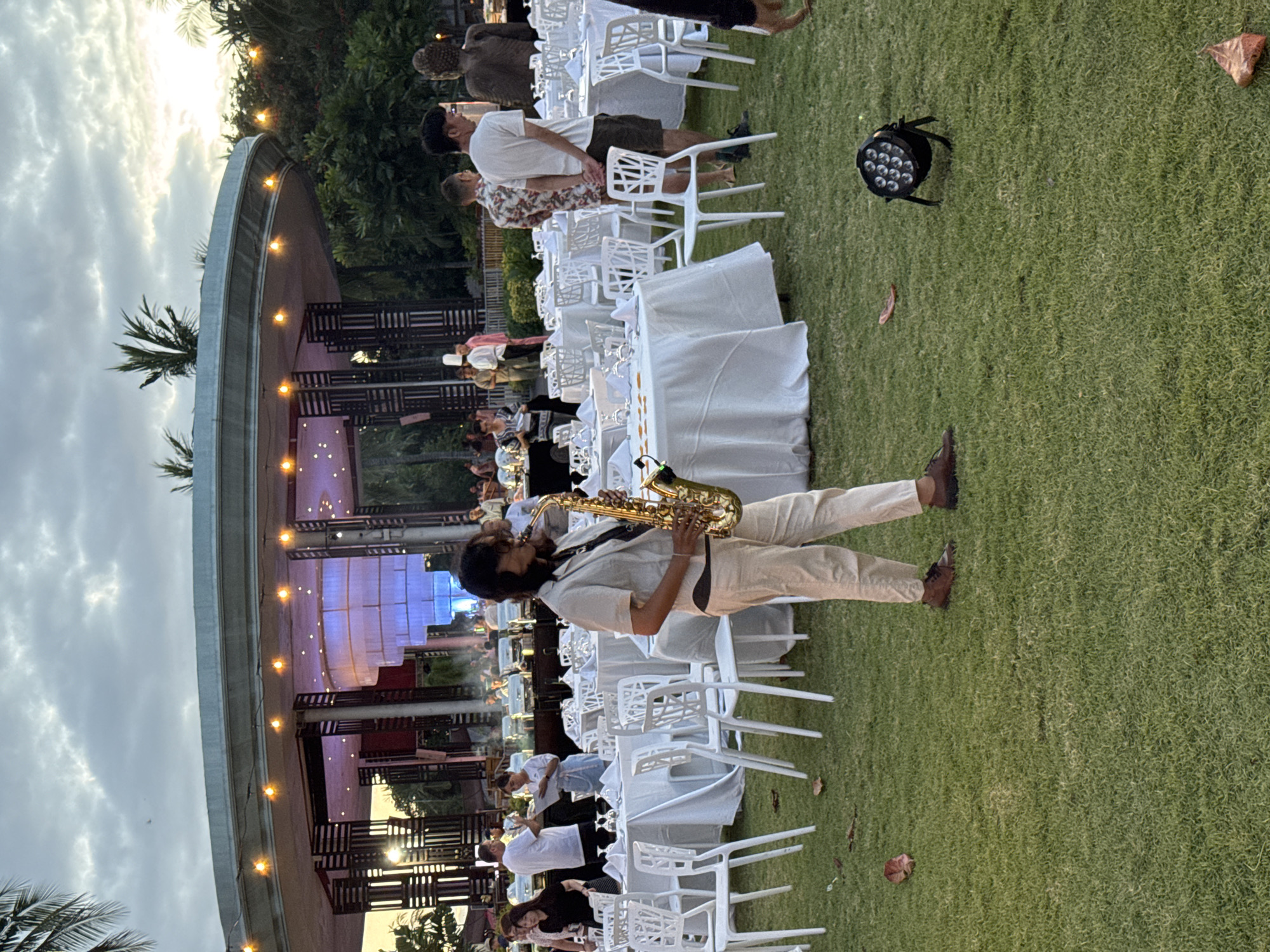 A wide-angle, outdoor shot shows a live jazz performance taking place in Kuta Selatan, Indonesia. A saxophonist, in cream-colored attire, stands on green grass and plays. Rows of white tables and chairs extend from behind him towards the viewer. A small crowd, largely in light-colored clothing, is visible in the background. A structure with a curved roof, outlined by lights, partially encloses the seating. The sky is overcast with clouds. A spotlight is visible on the lawn to the right.